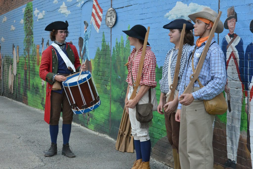 Four people dressed in historical costumes stand in front of a painted mural, with one person holding a drum and others holding rifles.