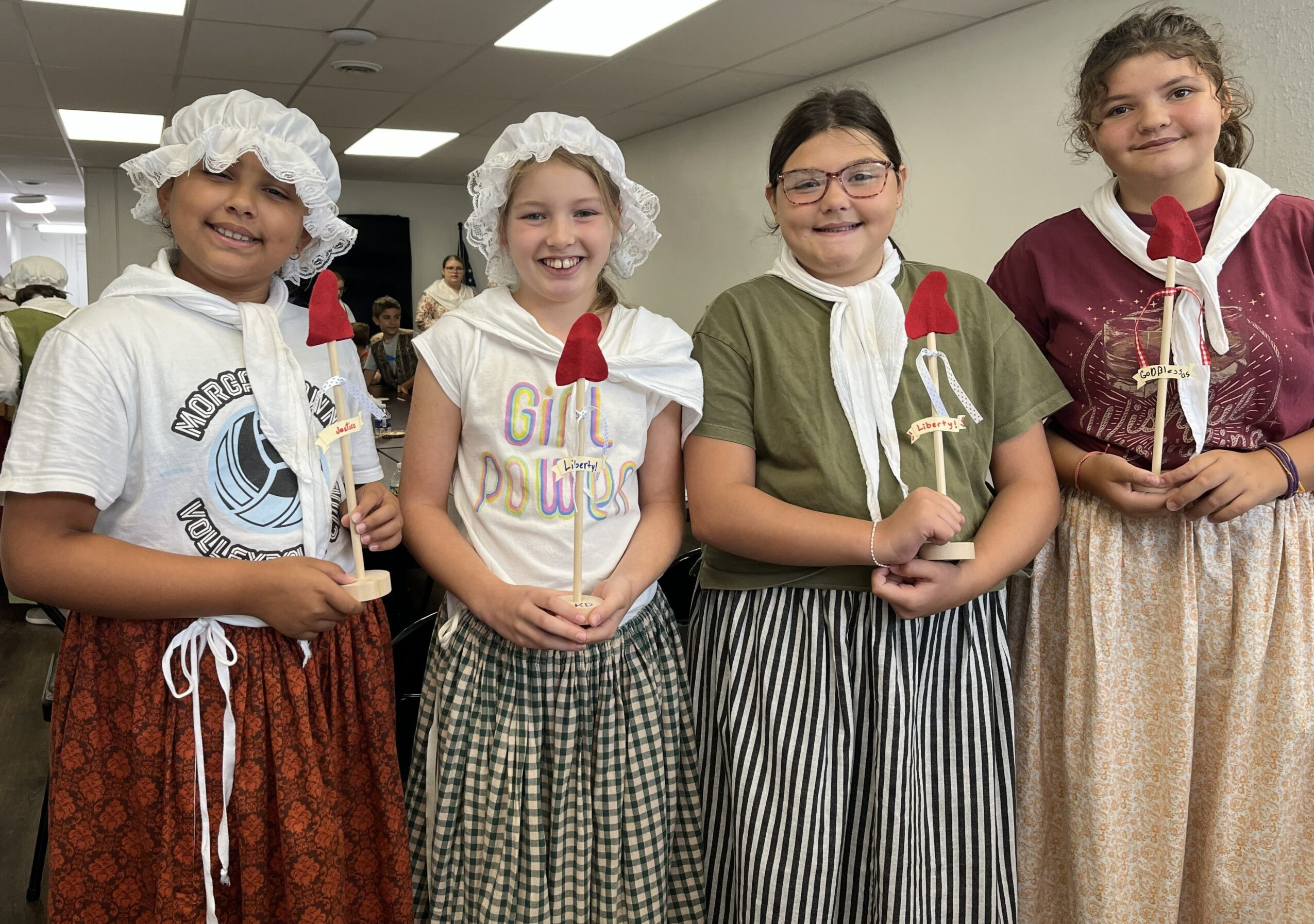 Five children in historical costumes holding small decorative objects, smiling indoors. Four girls wear bonnets and striped skirts, one girl wears glasses.