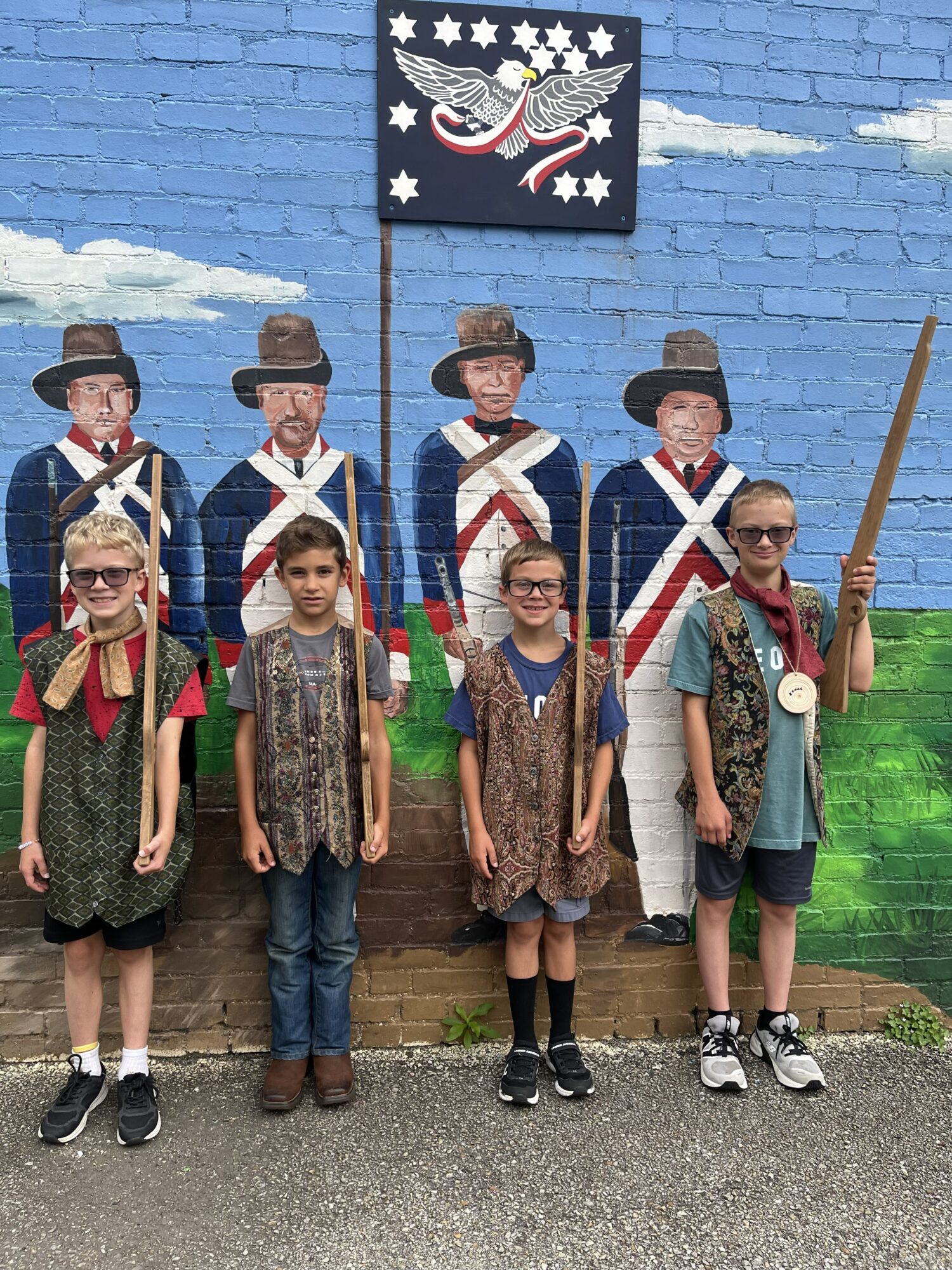 Group of children standing in front of a blue brick wall with a patriotic mural, some wearing costumes and glasses.