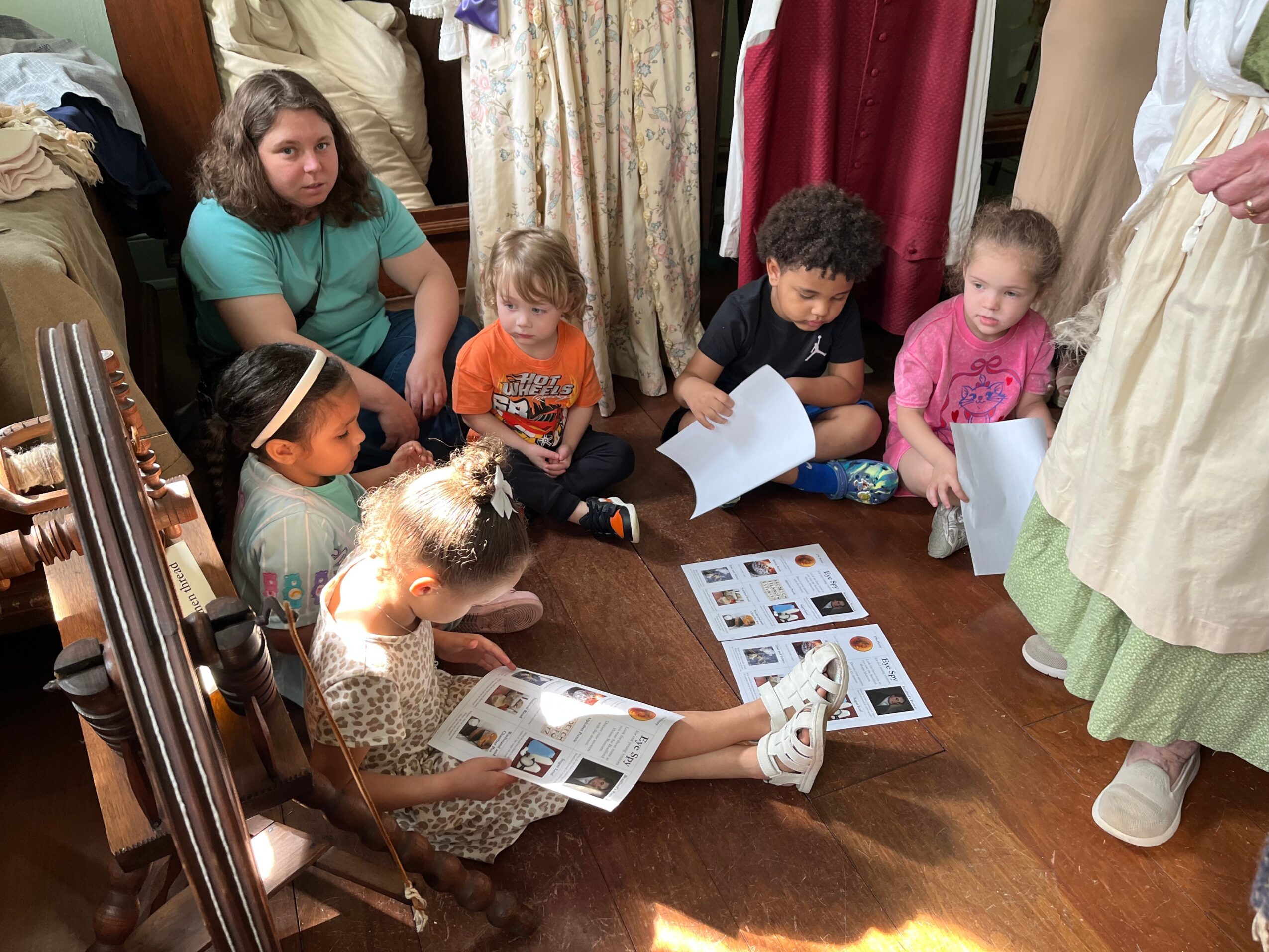 Group of children sitting on the floor, looking at papers and a person standing nearby, in a room with curtains and furniture.
