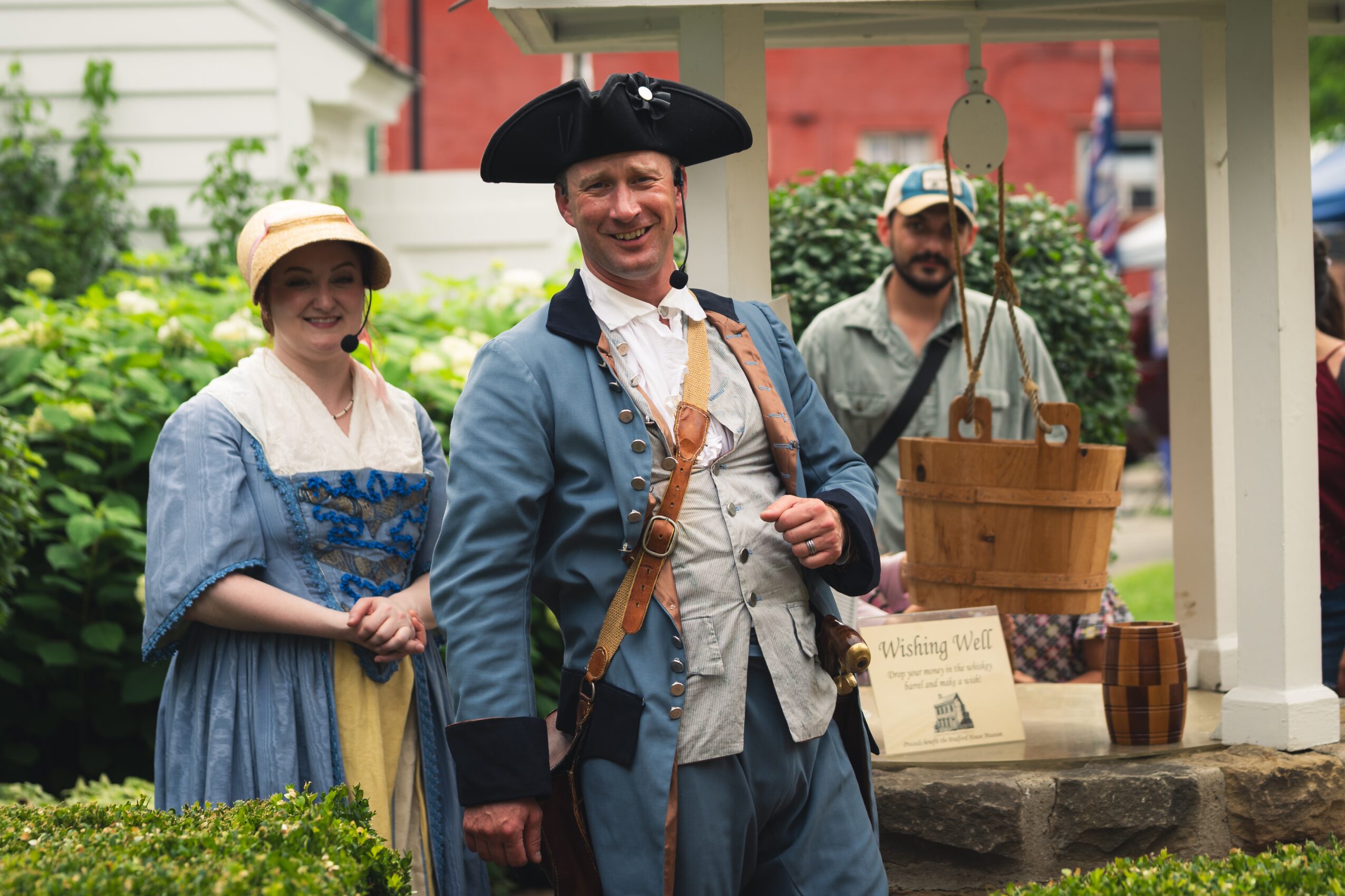 Three people dressed in historical clothing stand outdoors near a wooden structure, smiling at the camera.