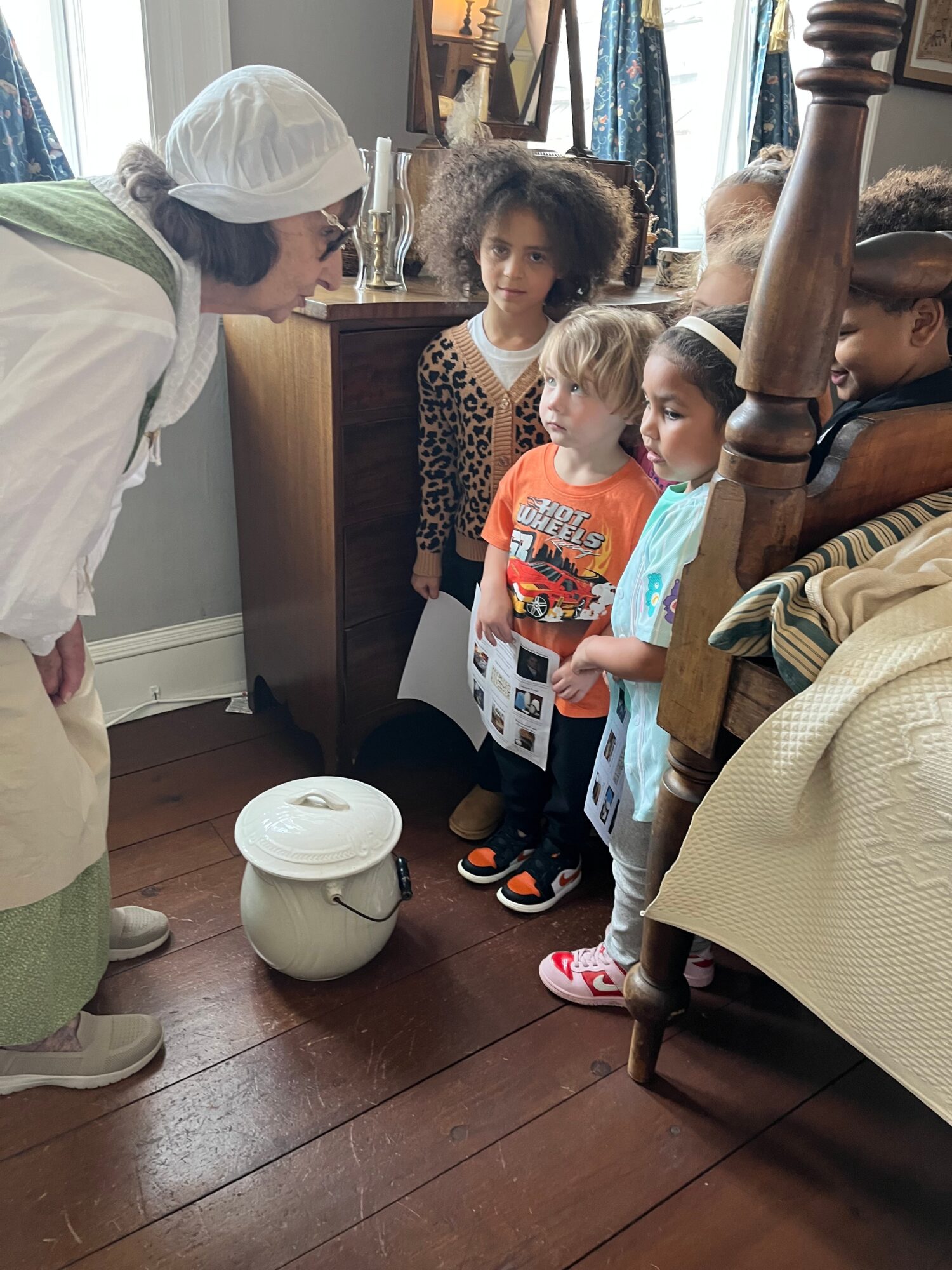 Adult woman with a hairnet talking to four children in a room with wooden furniture and a white device on the floor.