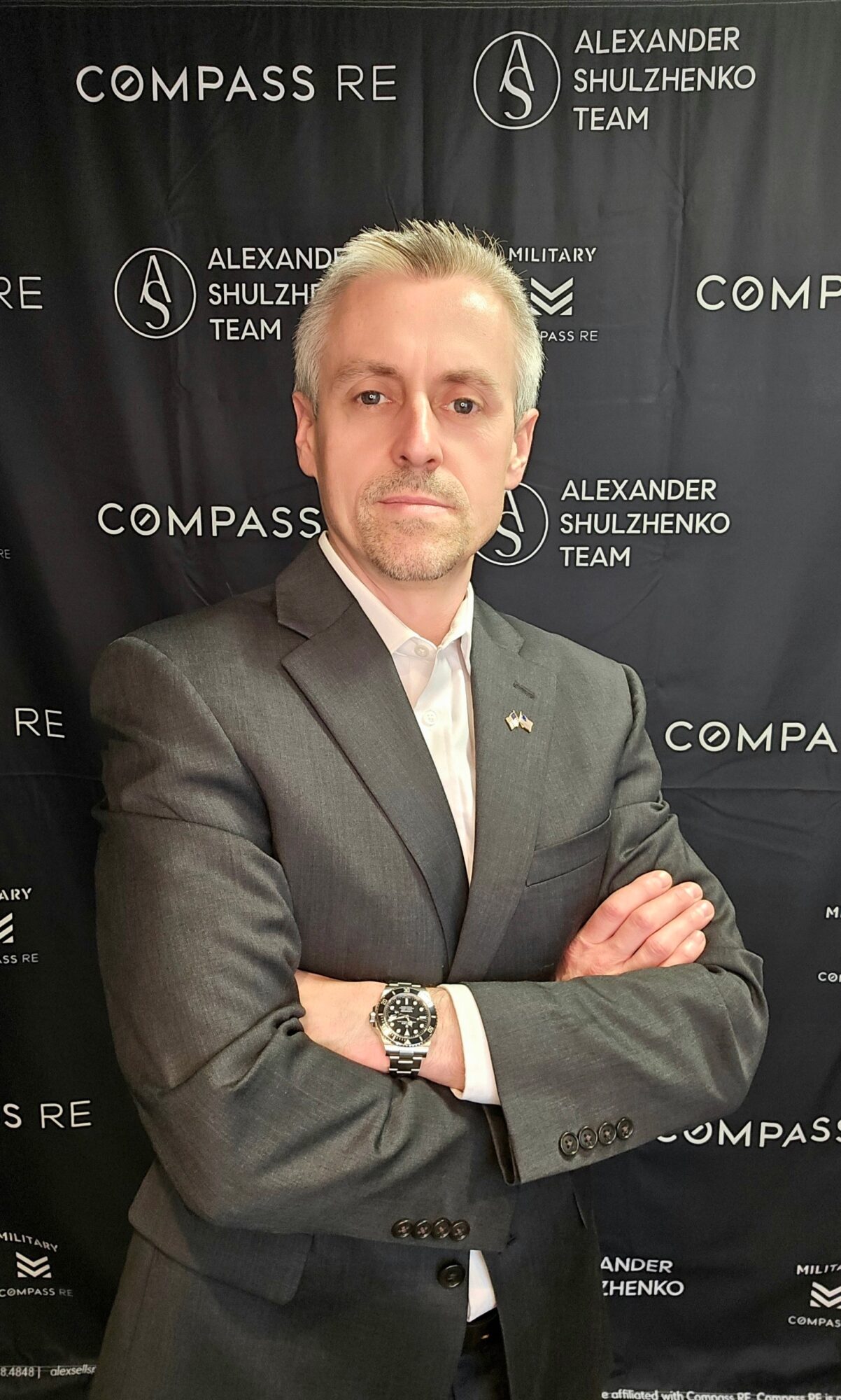 Man in suit with crossed arms standing in front of a black backdrop with logos and text.