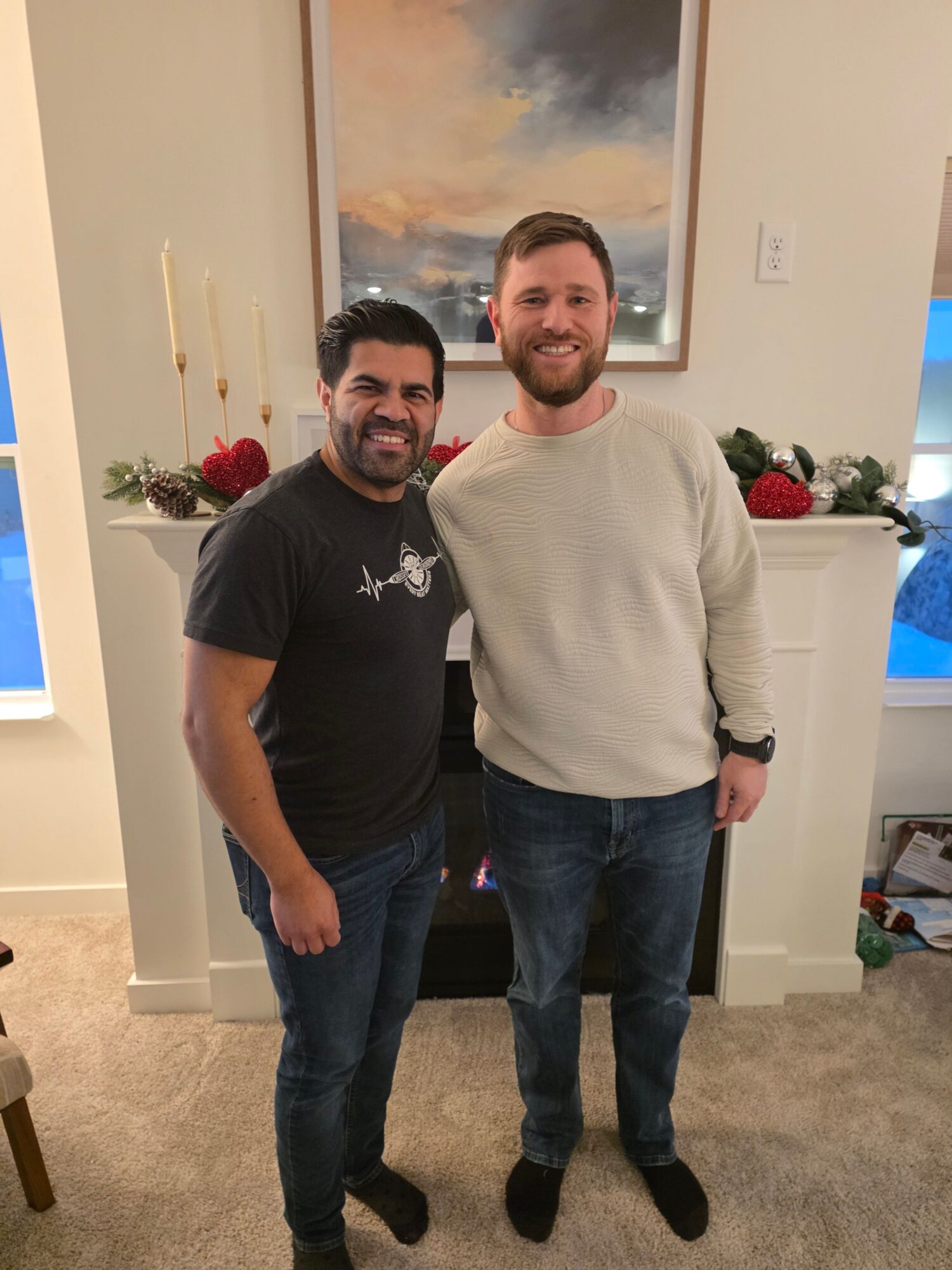Two men standing indoors in front of a white fireplace with Christmas decorations, smiling at the camera.