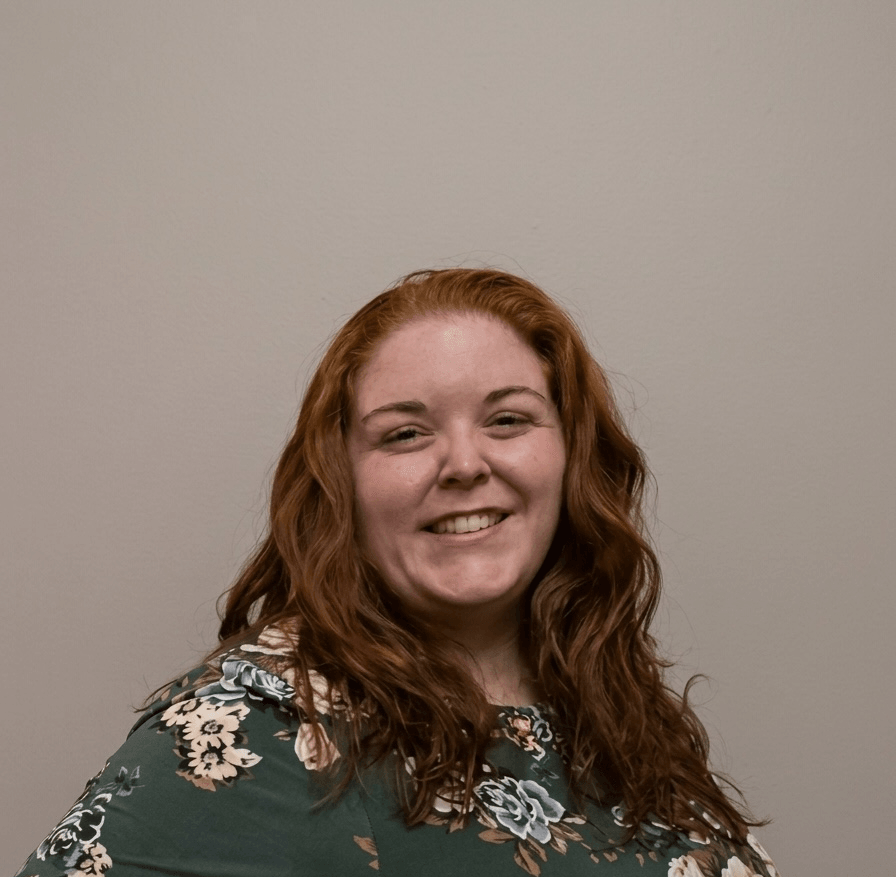 Smiling woman with long, wavy red hair wearing a floral top, standing against a plain wall.