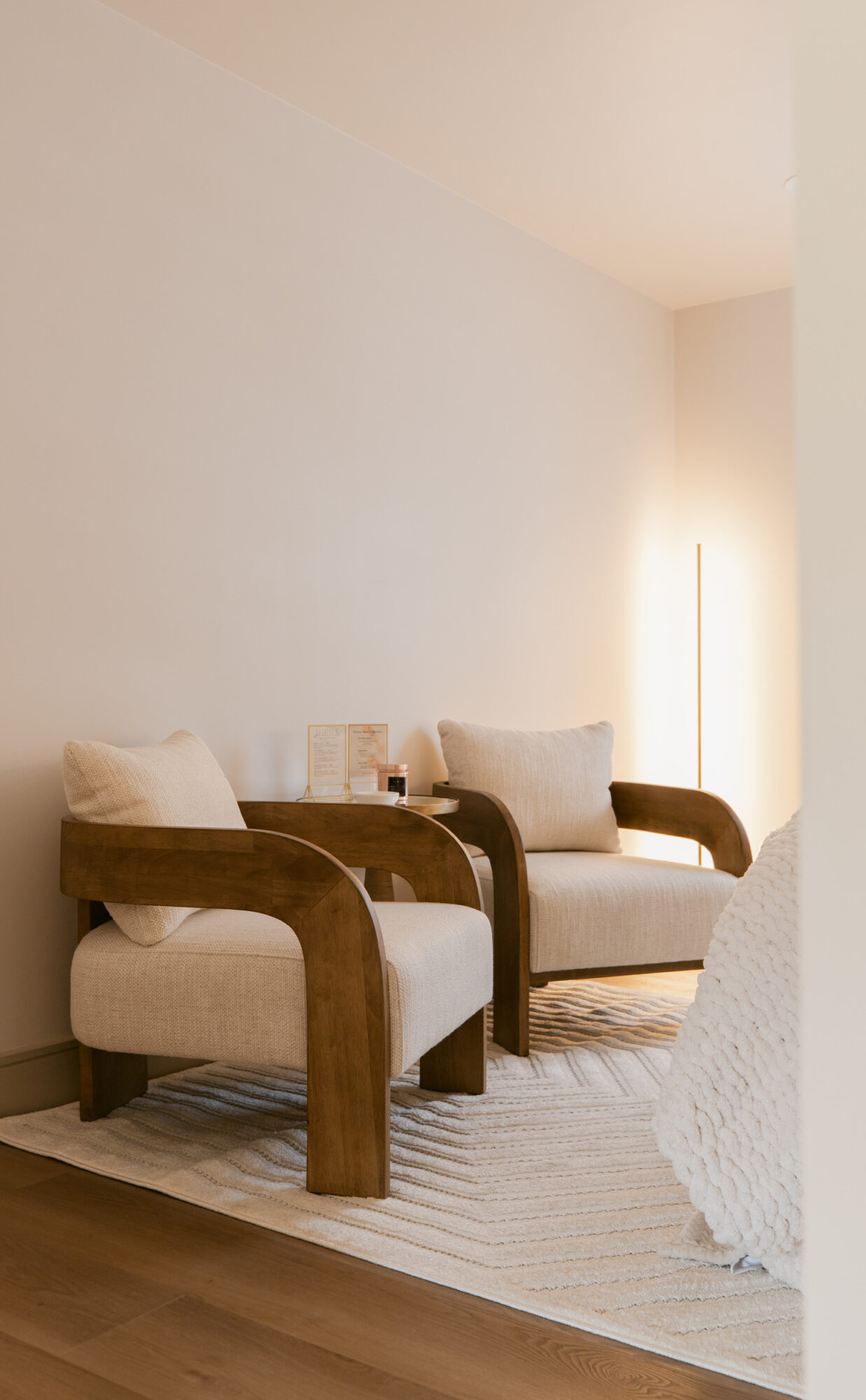 Two beige armchairs with wooden arms and cushions in a bright room, with a white rug and a light-colored wall.