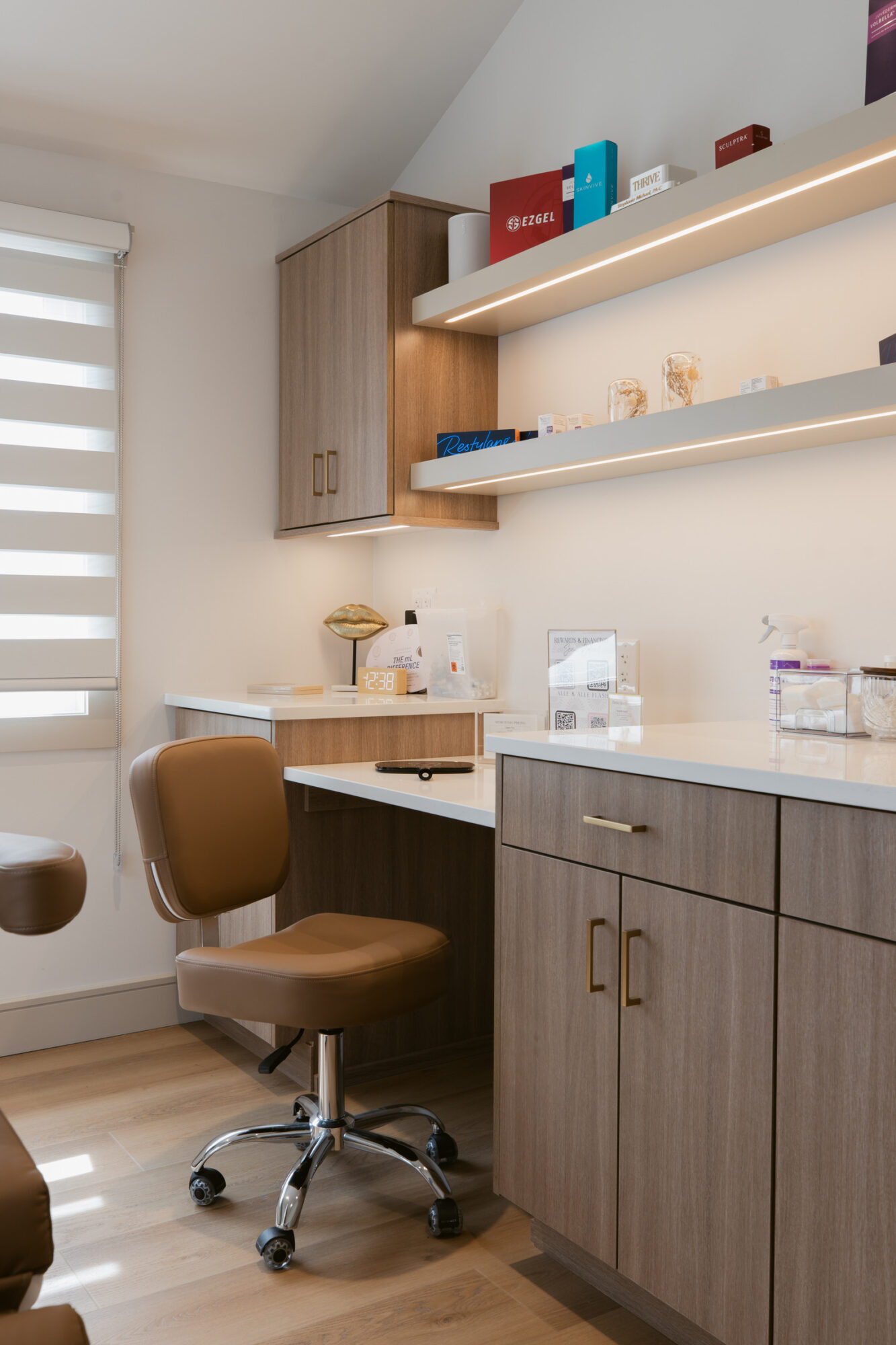Home office corner with desk, chair, shelves, and storage cabinets, illuminated by natural light from window with blinds.