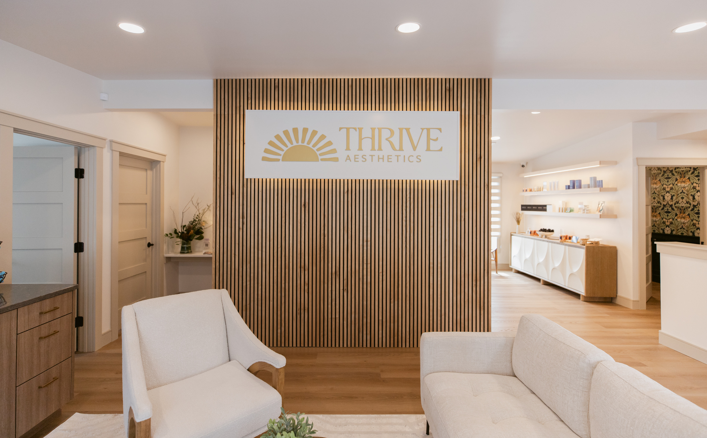 Reception area with white chairs, wooden wall, and a sign reading 'Thrive Esthetics' in a bright, modern space.