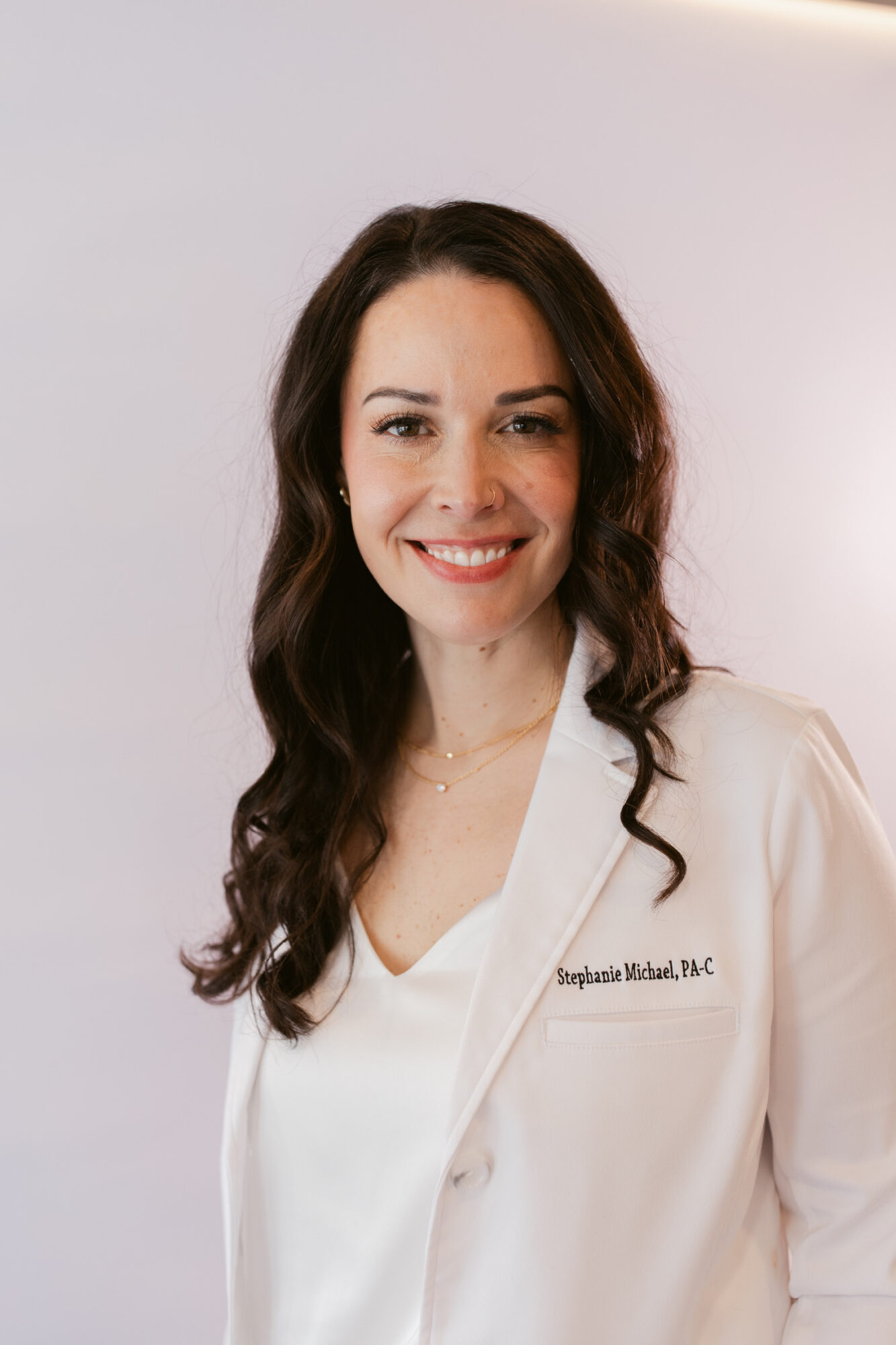Smiling woman with long dark hair in a white medical coat, standing against a plain background.