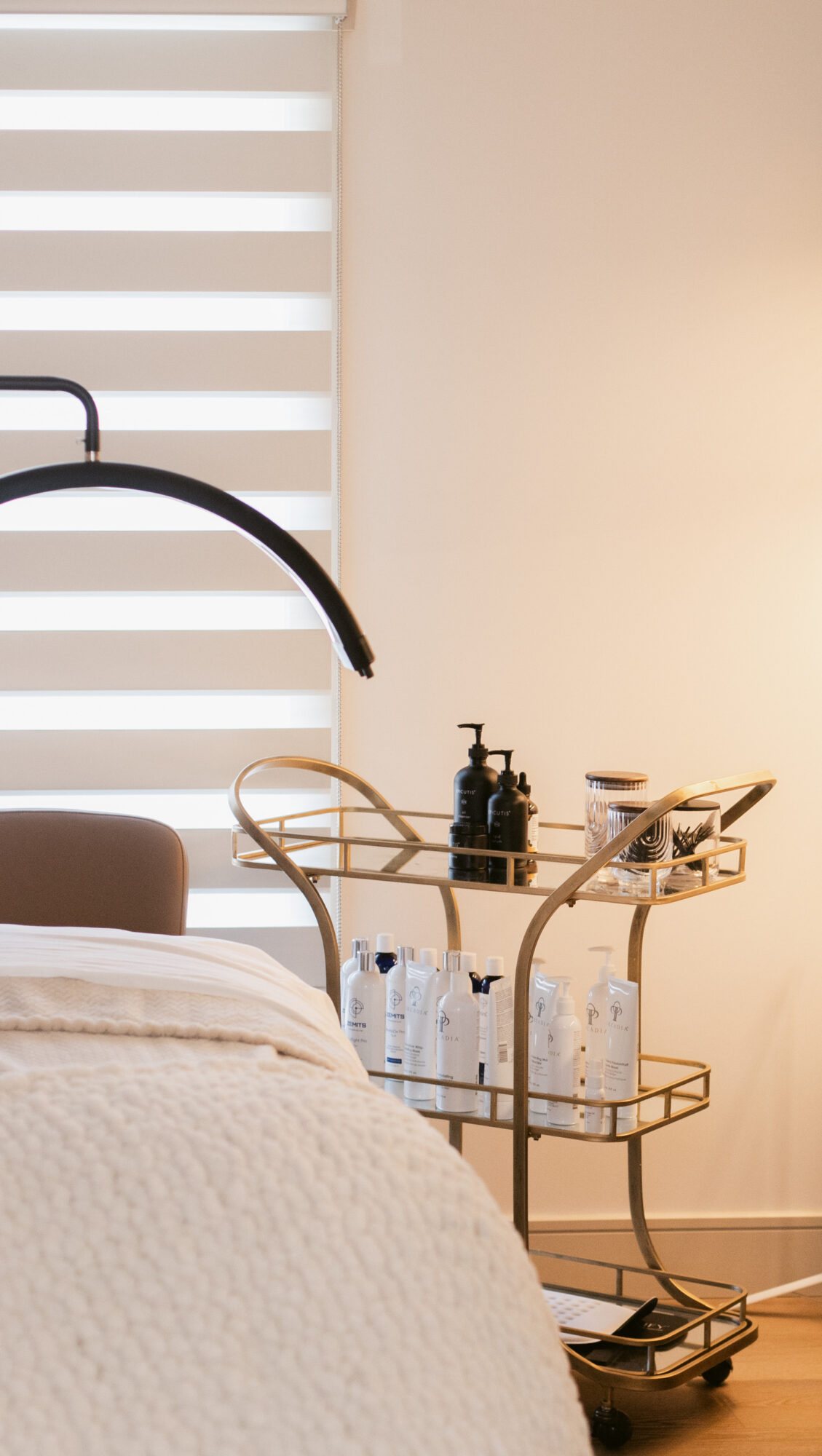 Hospital bed with beige bedding and a metal bedside cart holding bottles and medical supplies, near a window with blinds.
