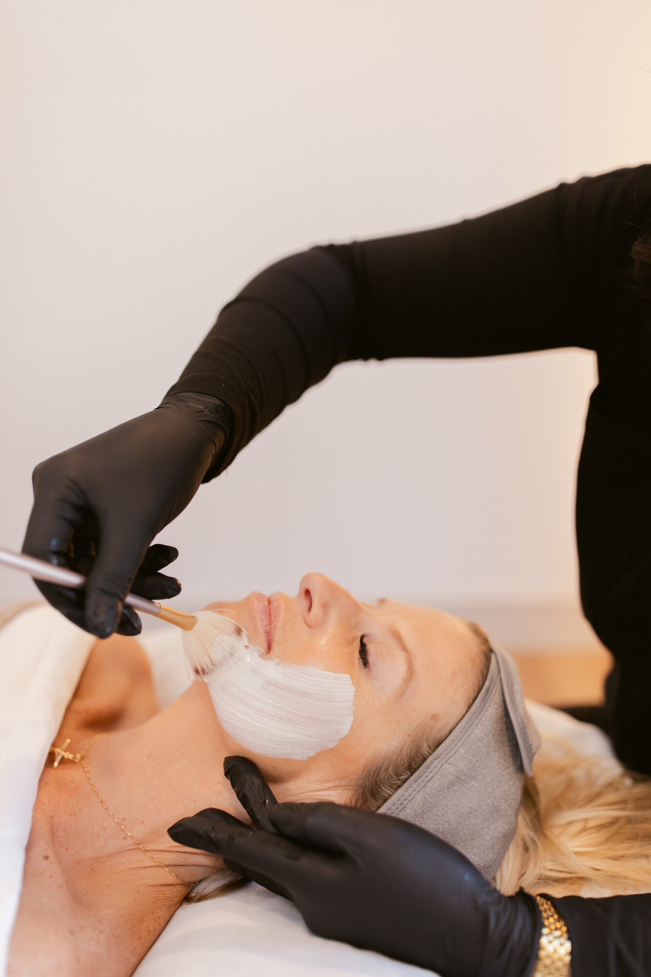 Person receiving a cosmetic treatment on face, lying down with head supported, practitioner wearing black gloves applying product.