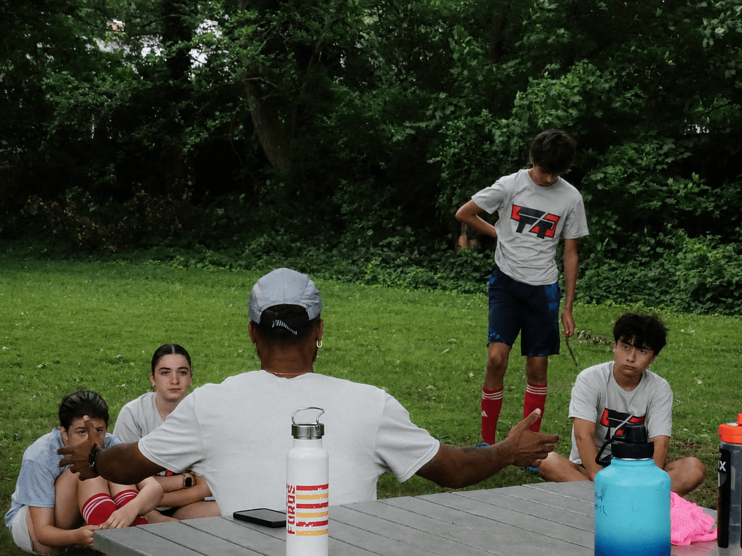Group of children and an adult outdoors on grass with trees in background, some sitting and one standing, engaging in activity.