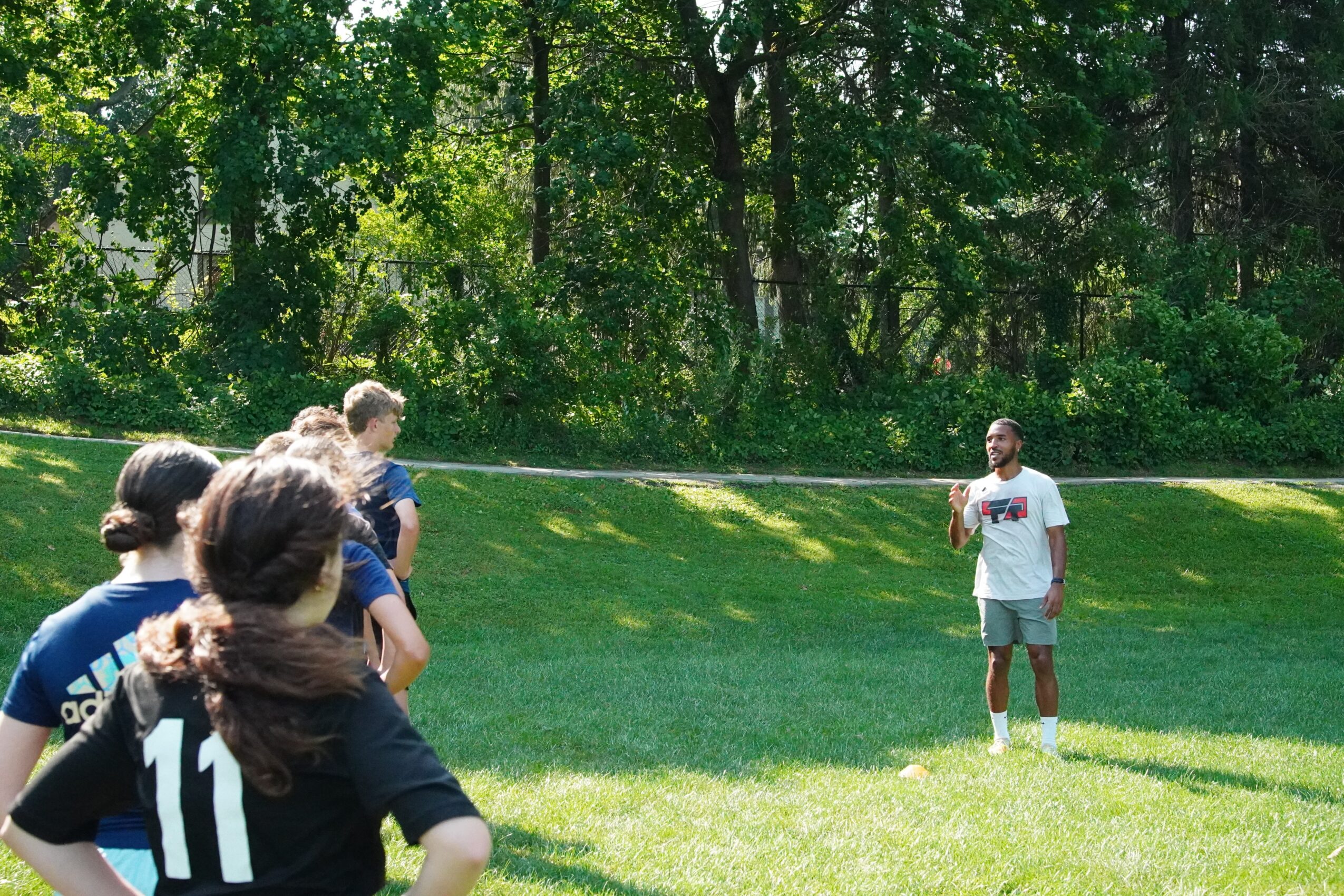 Group of children standing on grass in a park, facing a person holding a microphone, with trees in the background.