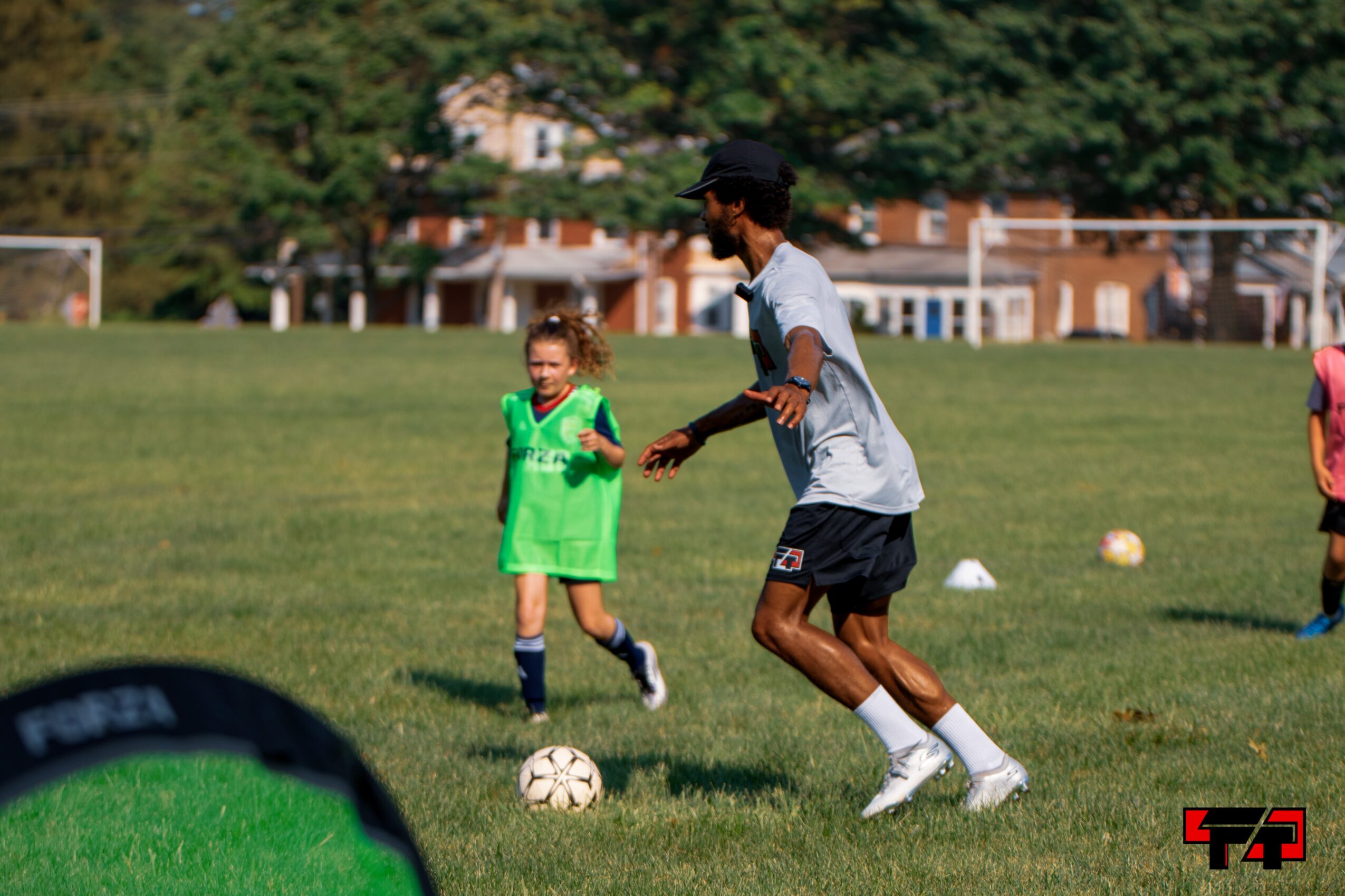 A young girl in a green sports uniform runs on a grassy field, with a man in a gray shirt and black shorts nearby, playing soccer.