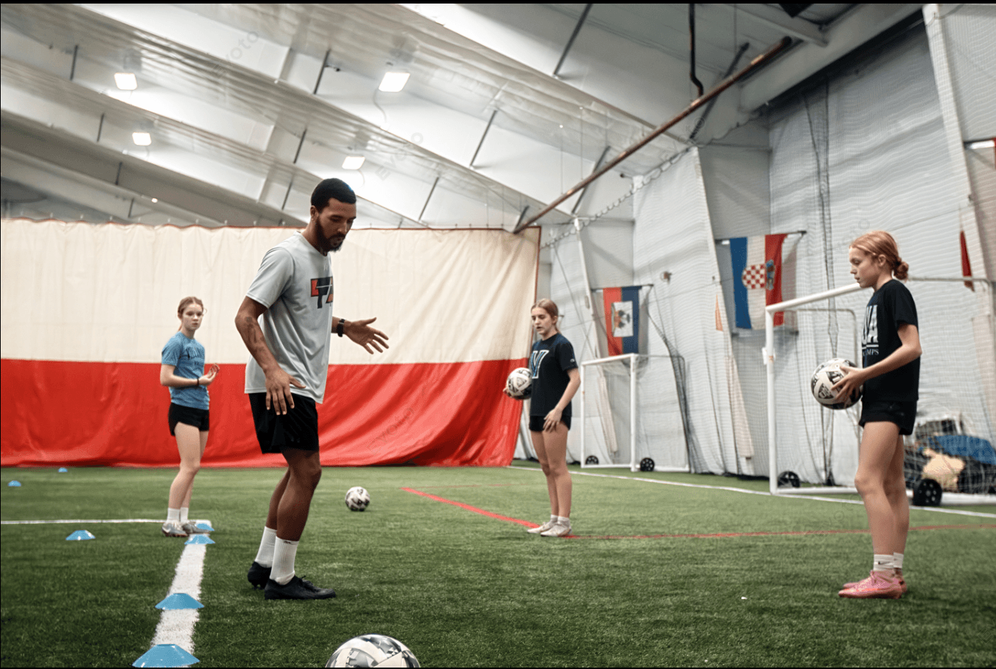 Coach instructs four children holding soccer balls in an indoor sports facility with Croatian flags on the wall.