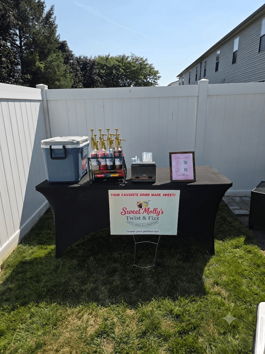 Table with drinks, a cooler, and a framed sign outdoors on grass, against a white fence and house.