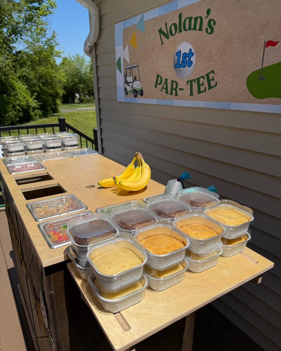 Table with bananas and containers of food outside near a building with a sign reading 'Nolan's 1st PAR-TEE'.