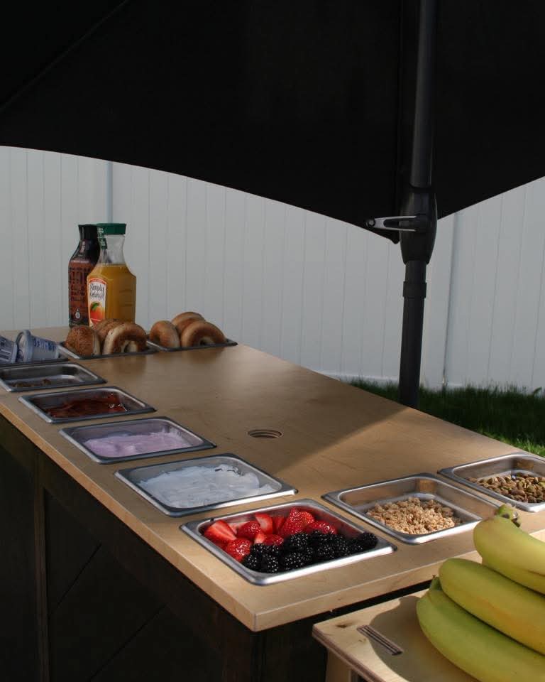 Table with food containers, bananas, and condiments under a black umbrella, outdoor setting with a white fence in background.