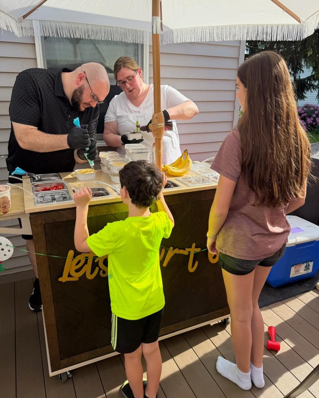 Two children stand in front of a food stand with two adults preparing food behind the counter, outdoors on a sunny day.