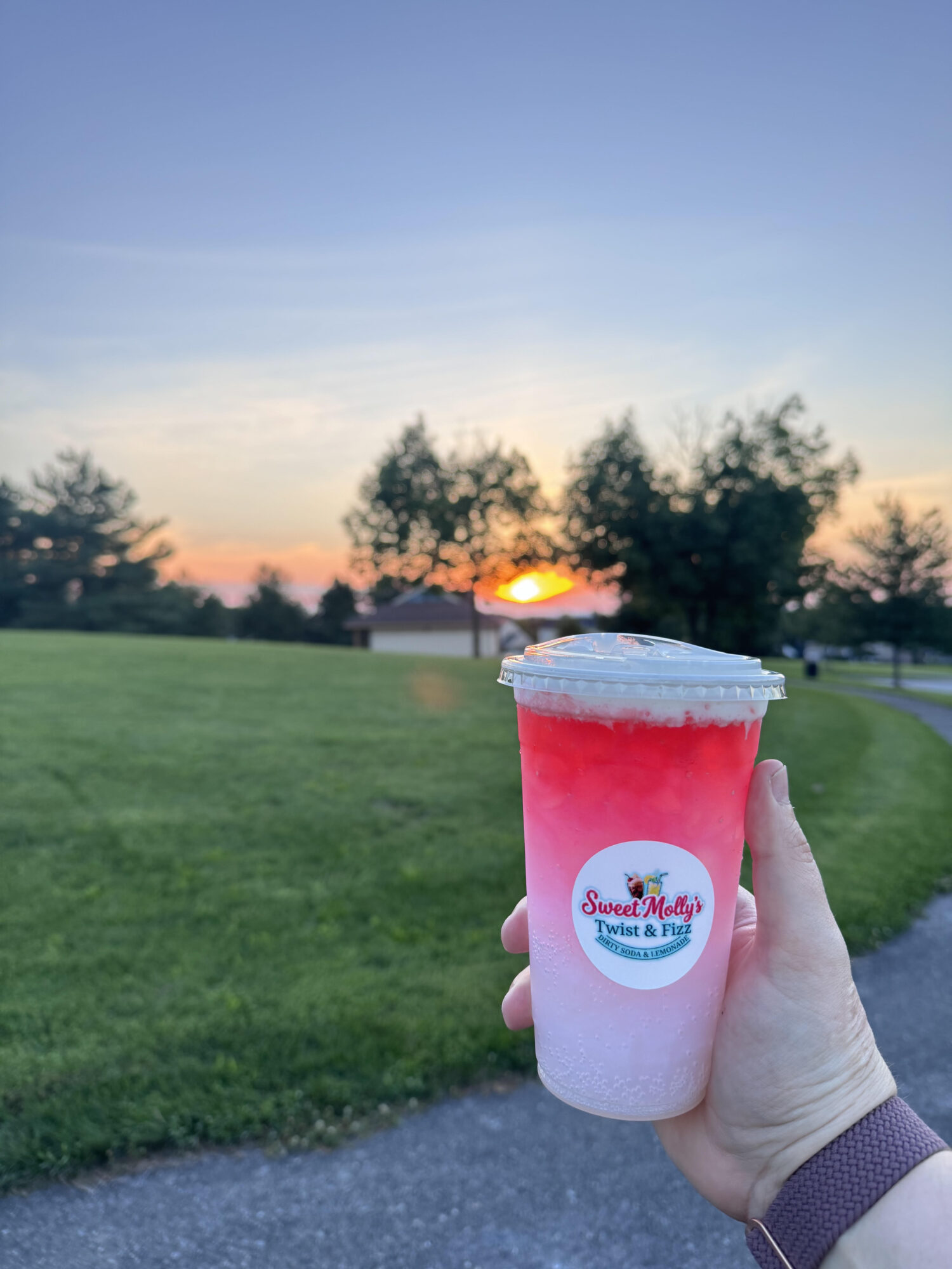 Person holding a pink and white drink with a lid outdoors during sunset, trees and a grassy field in background.