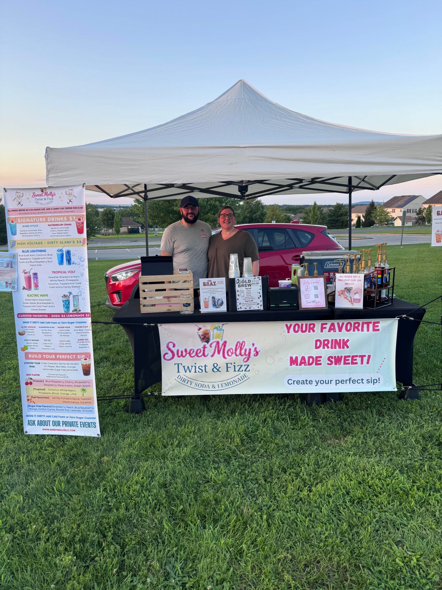 Stand with a tent and table displaying drinks and signs at an outdoor event, with a red vehicle in the background.