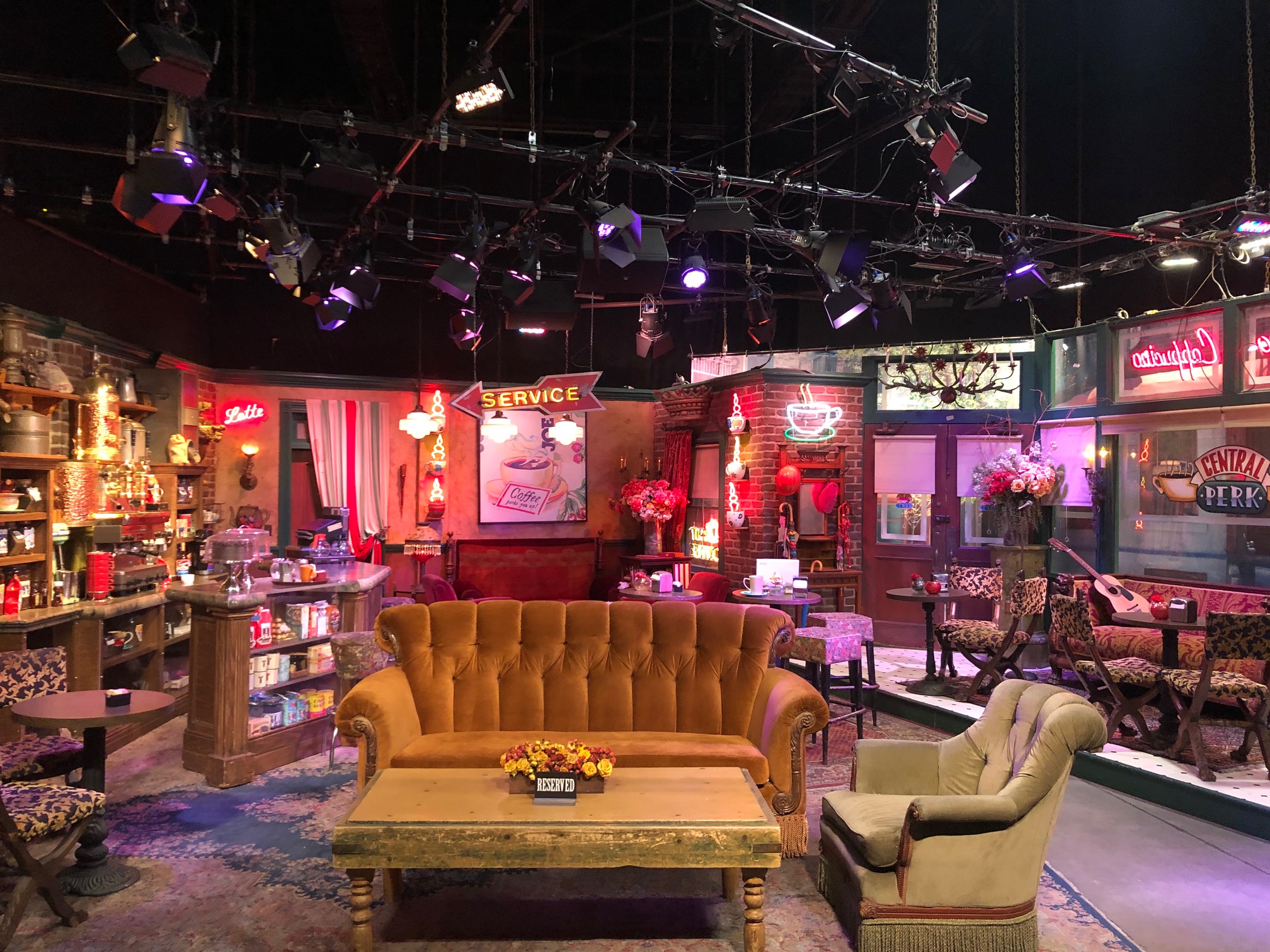 A cozy lounge with a mustard sofa, armchair, and a coffee table, decorated with pink flowers and vintage signs.