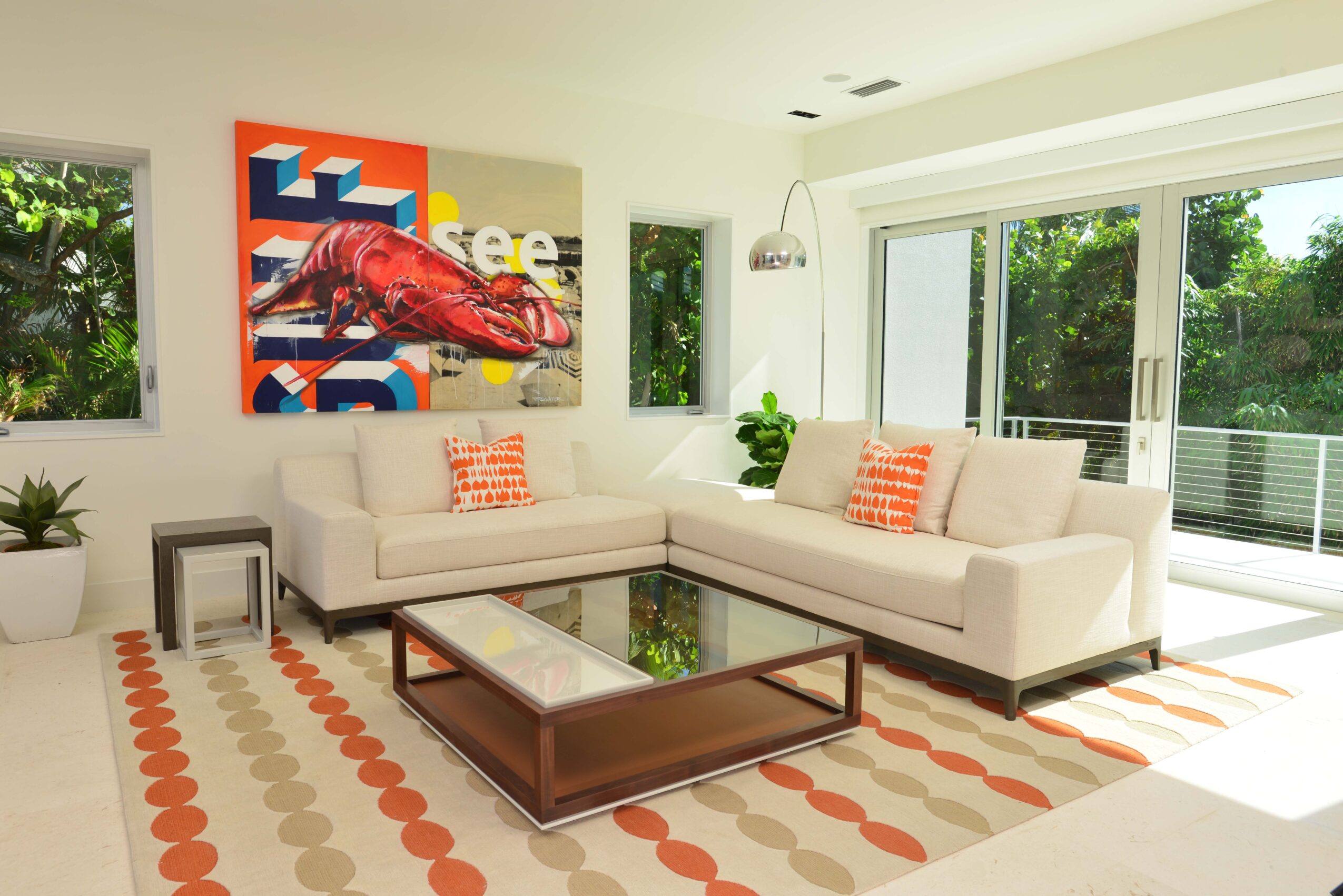 Living room with two white sofas, glass coffee table, colorful artwork, and large windows with greenery outside.