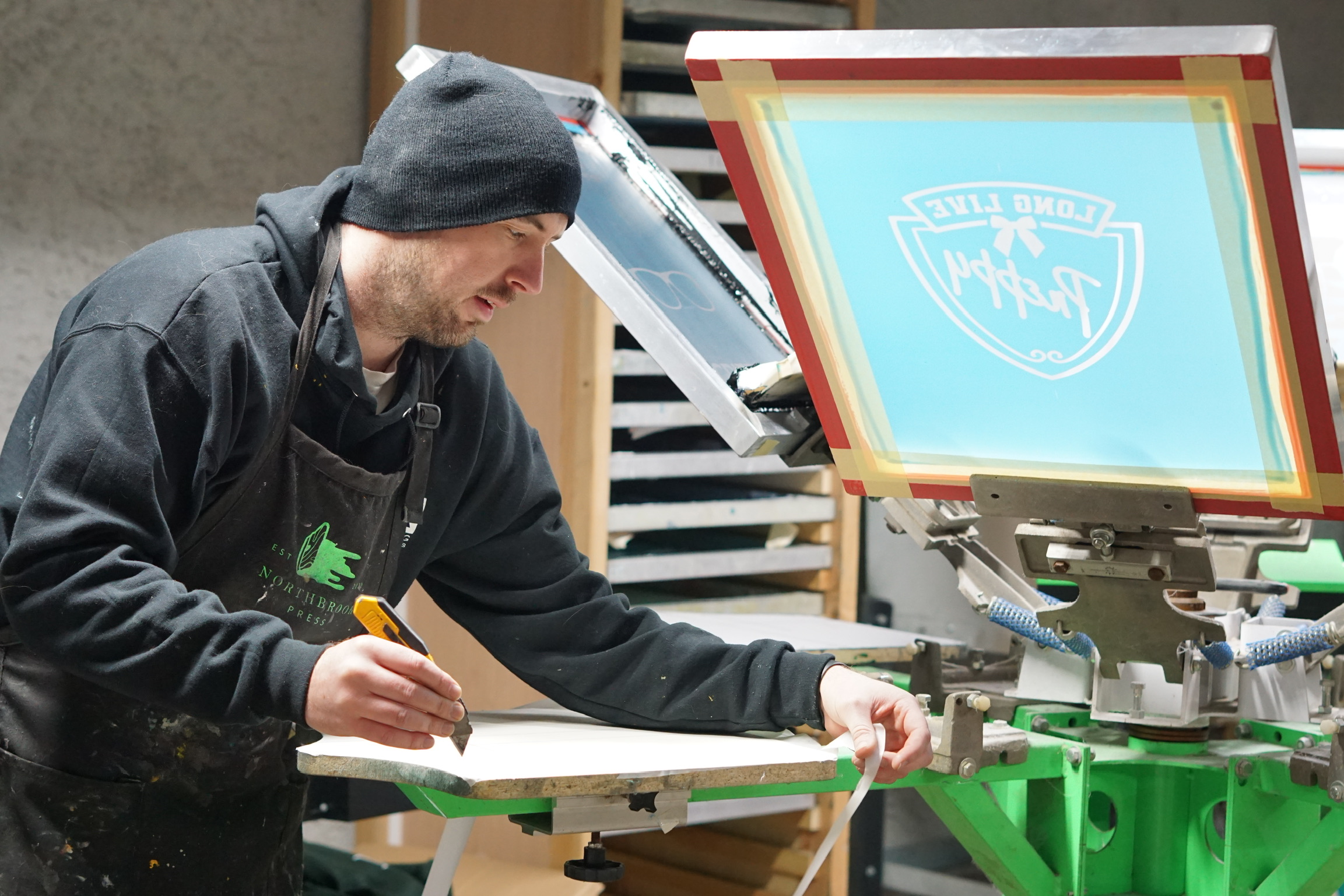 Person working with robotic arm and computer screen in workshop, writing on a notepad, wearing a hoodie and beanie.