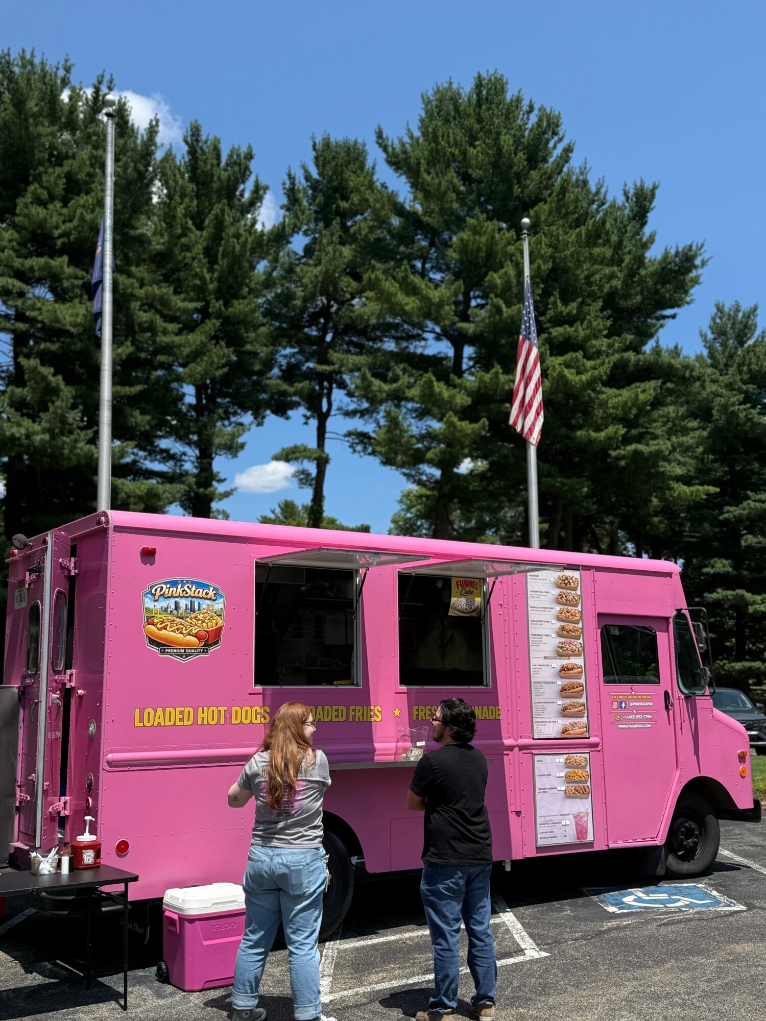 Pink food truck with two people standing in front, trees and an American flag in the background.