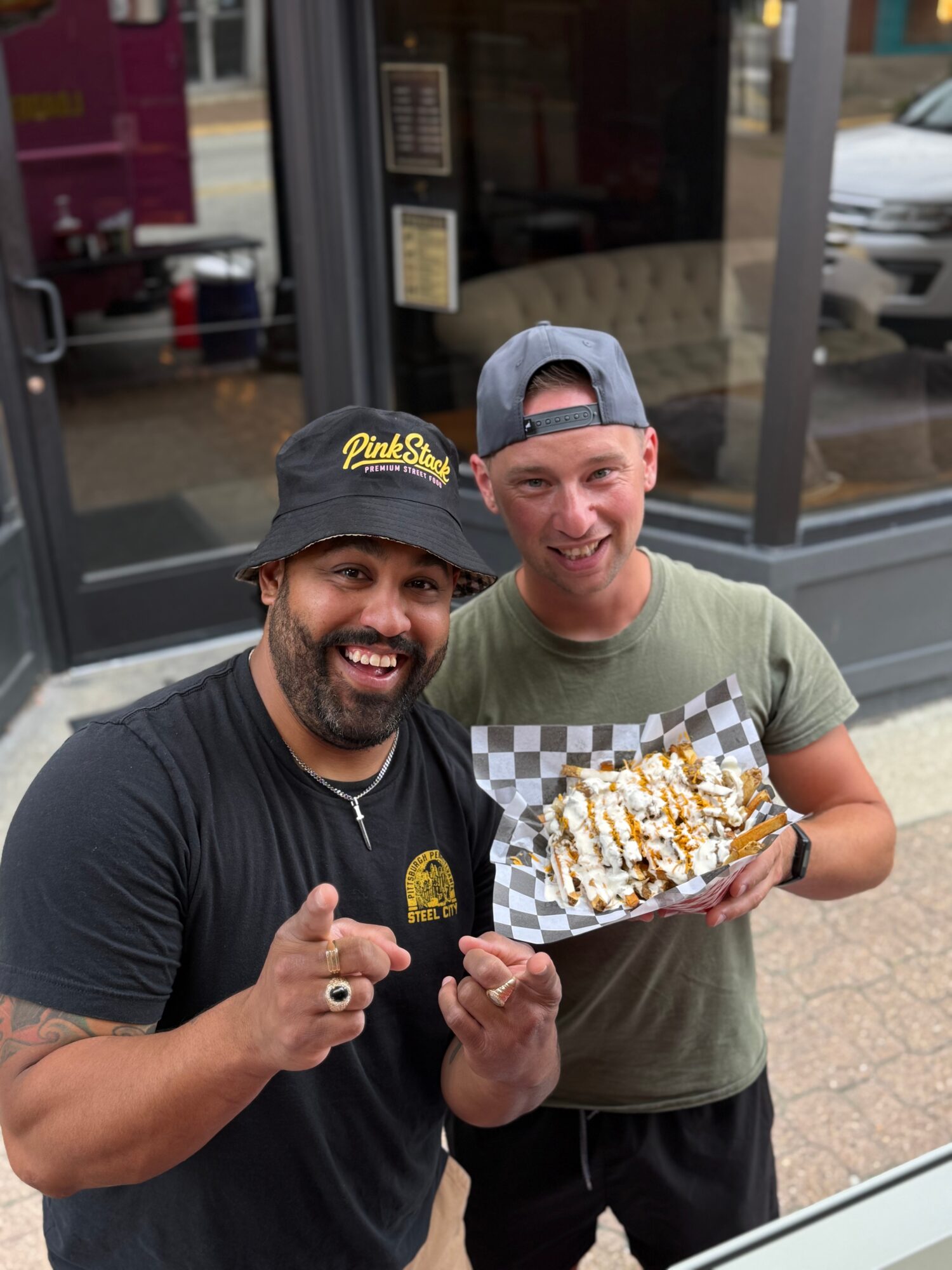 Two men smiling, one holding a tray of food with toppings, standing outside a store with glass windows.