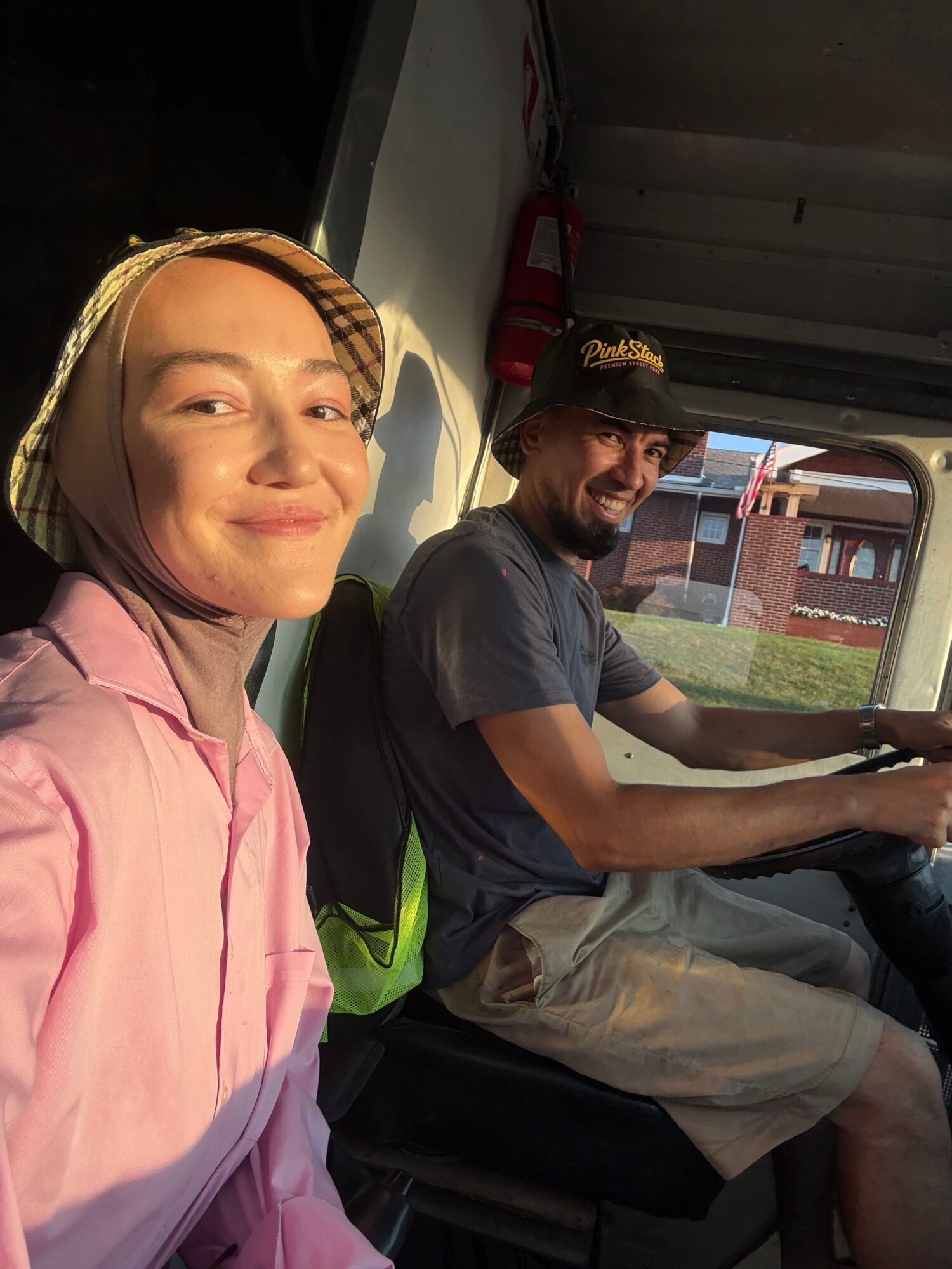 Two people sitting inside a vehicle, one in the foreground wearing a pink shirt and headscarf, the other in the background with a cap, looking outside.