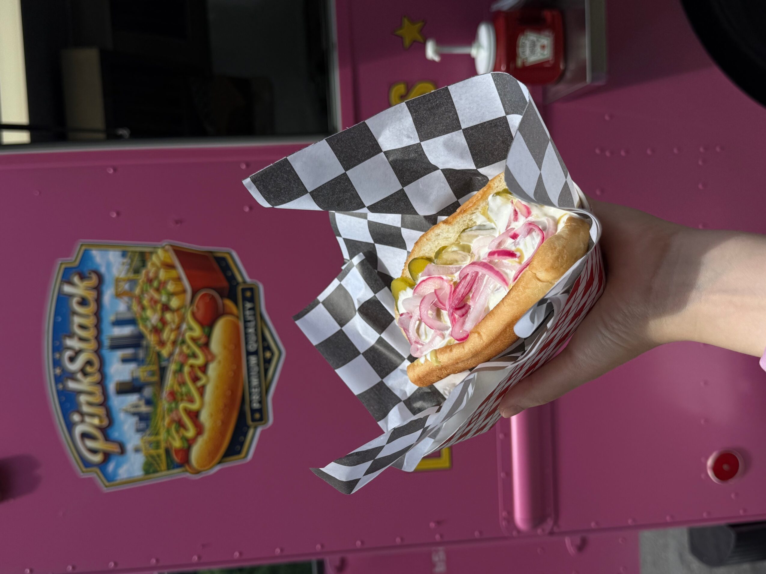 Hand holding a hot dog with pink and white toppings in checkered paper, pink background, PinStack sign visible.