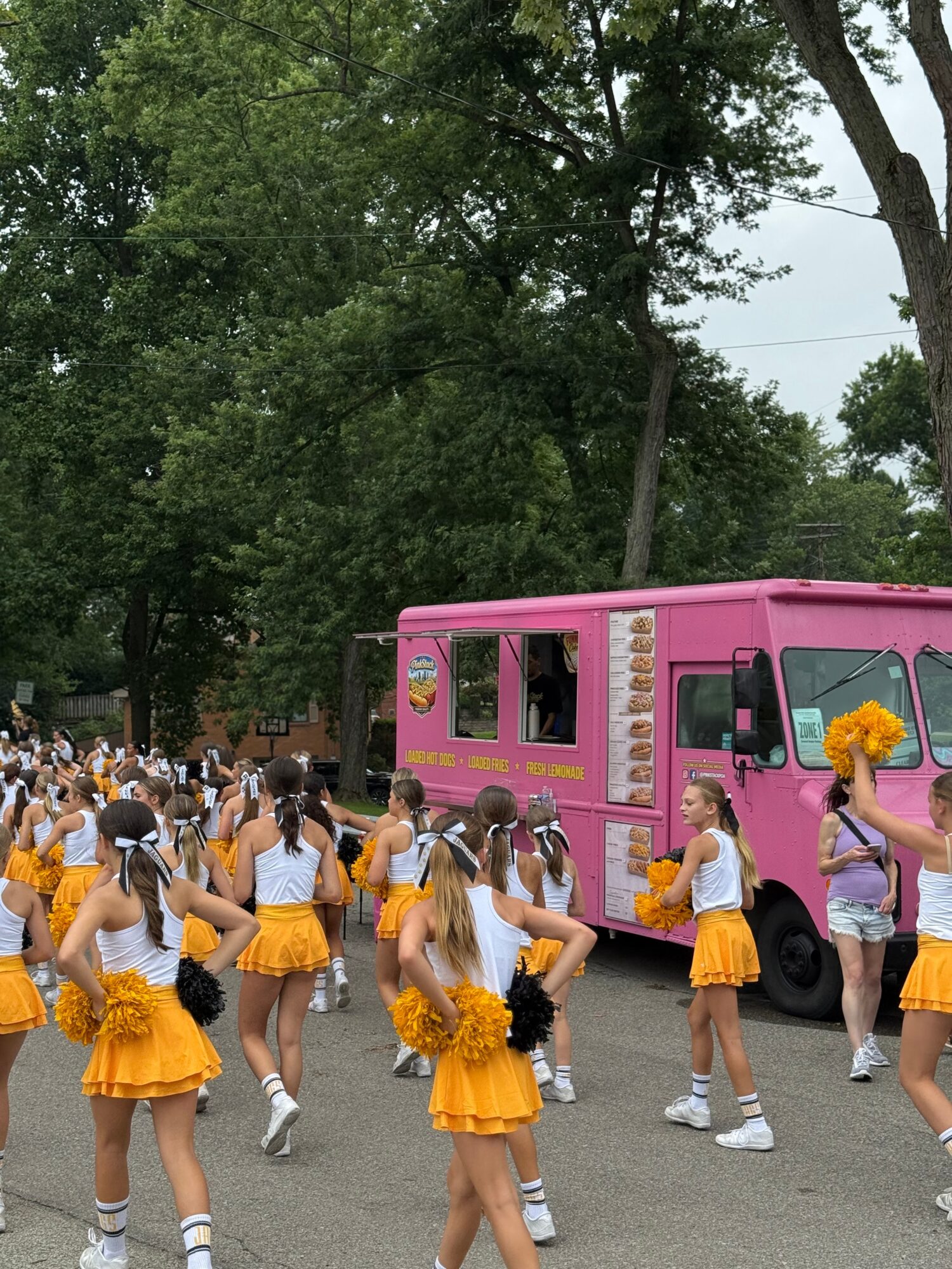 Group of cheerleaders in orange skirts and white tops standing outdoors near a pink vehicle, with trees in the background.
