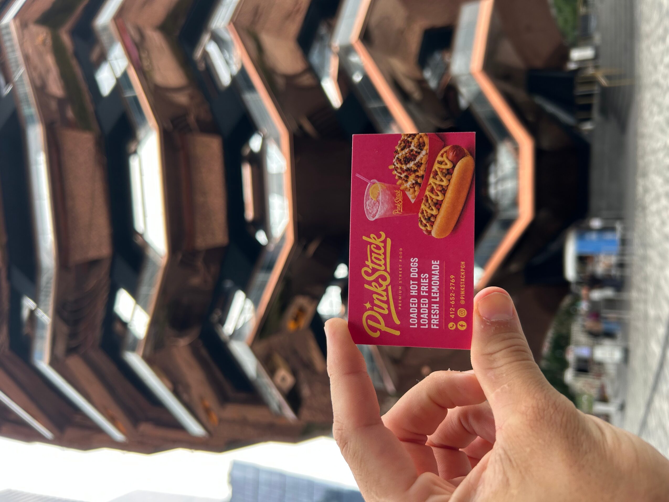 Hand holding a pink card with a hot dog illustration and text, in front of a store display with shelves and products.