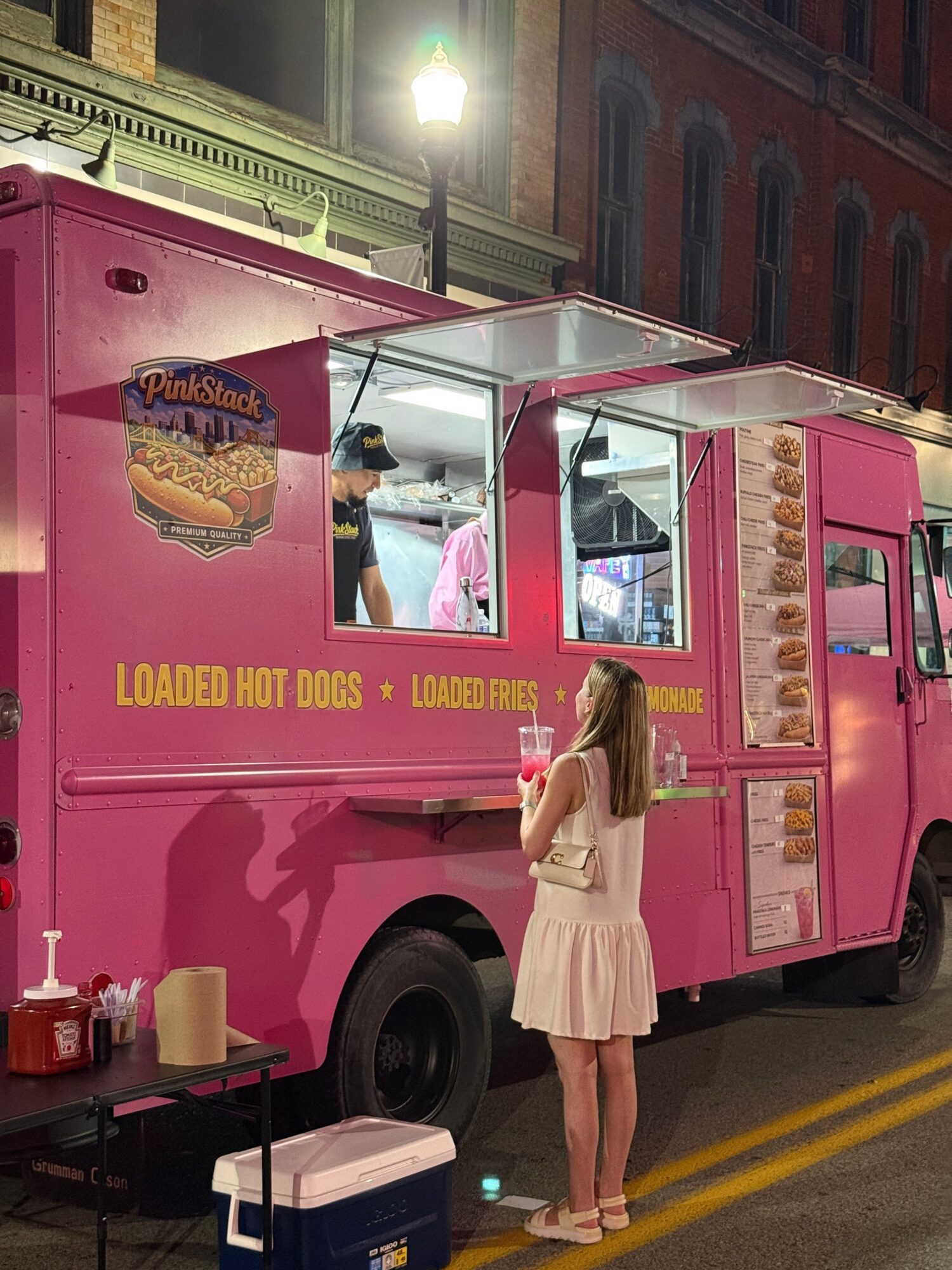 Young woman in light pink dress and heels stands next to a pink ice cream truck on a city street at night.