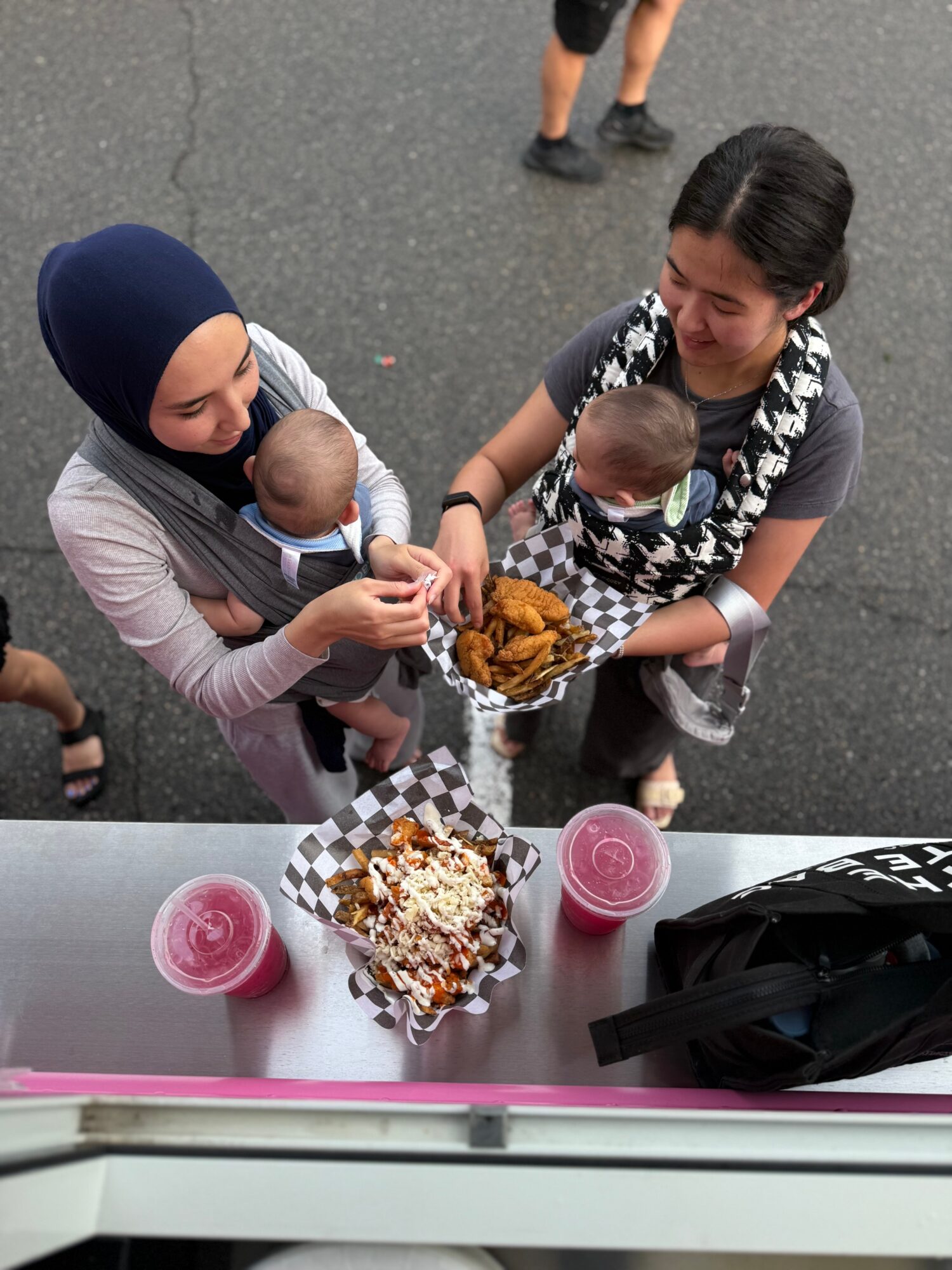 Two women holding babies and sharing food at an outdoor event, with food trays and drinks on a table.