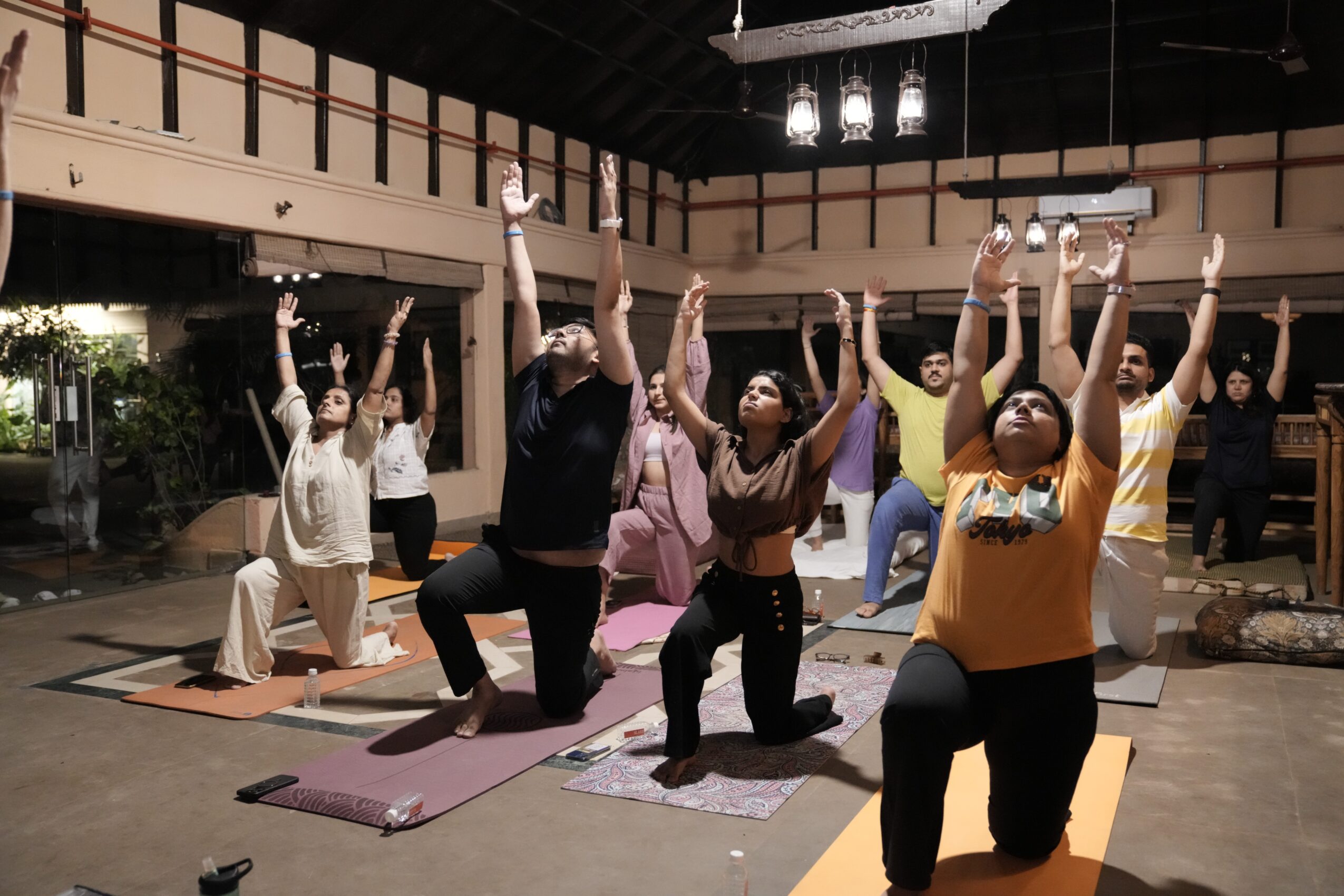 Group of people practicing yoga with arms raised indoors, some kneeling on mats, in a spacious room with high ceiling.