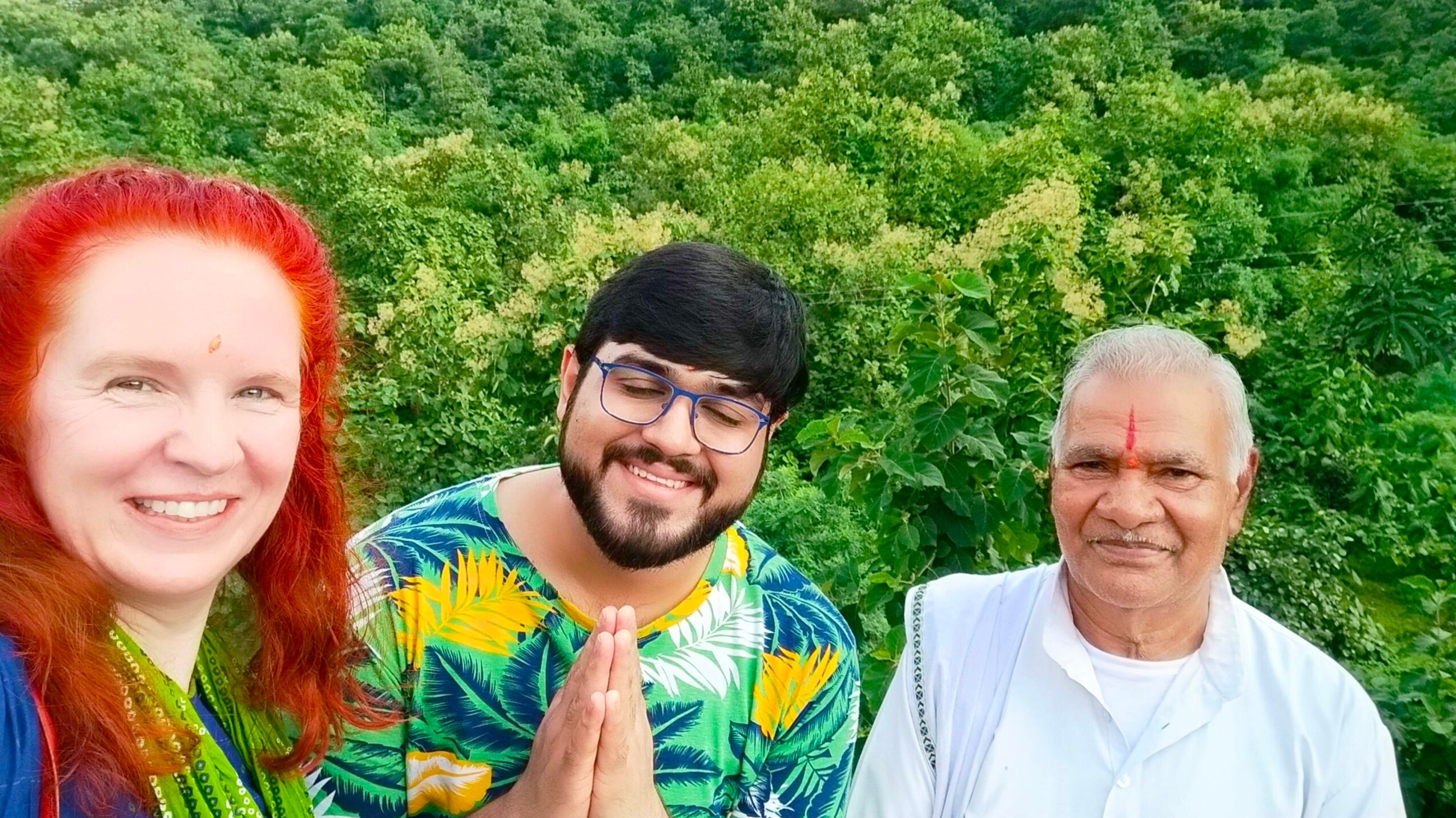 Three people outdoors with green trees in the background, smiling, one with hands in a prayer gesture, one with red hair, one with glasses.