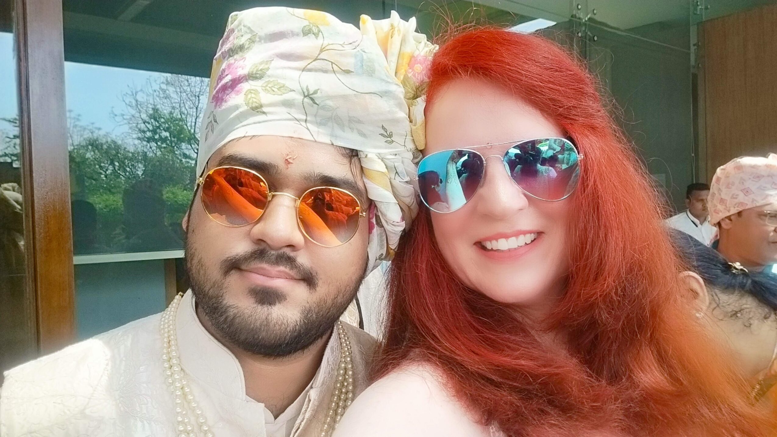 Two people smiling, one with red hair and sunglasses, the other with a headscarf and orange sunglasses, outdoors.