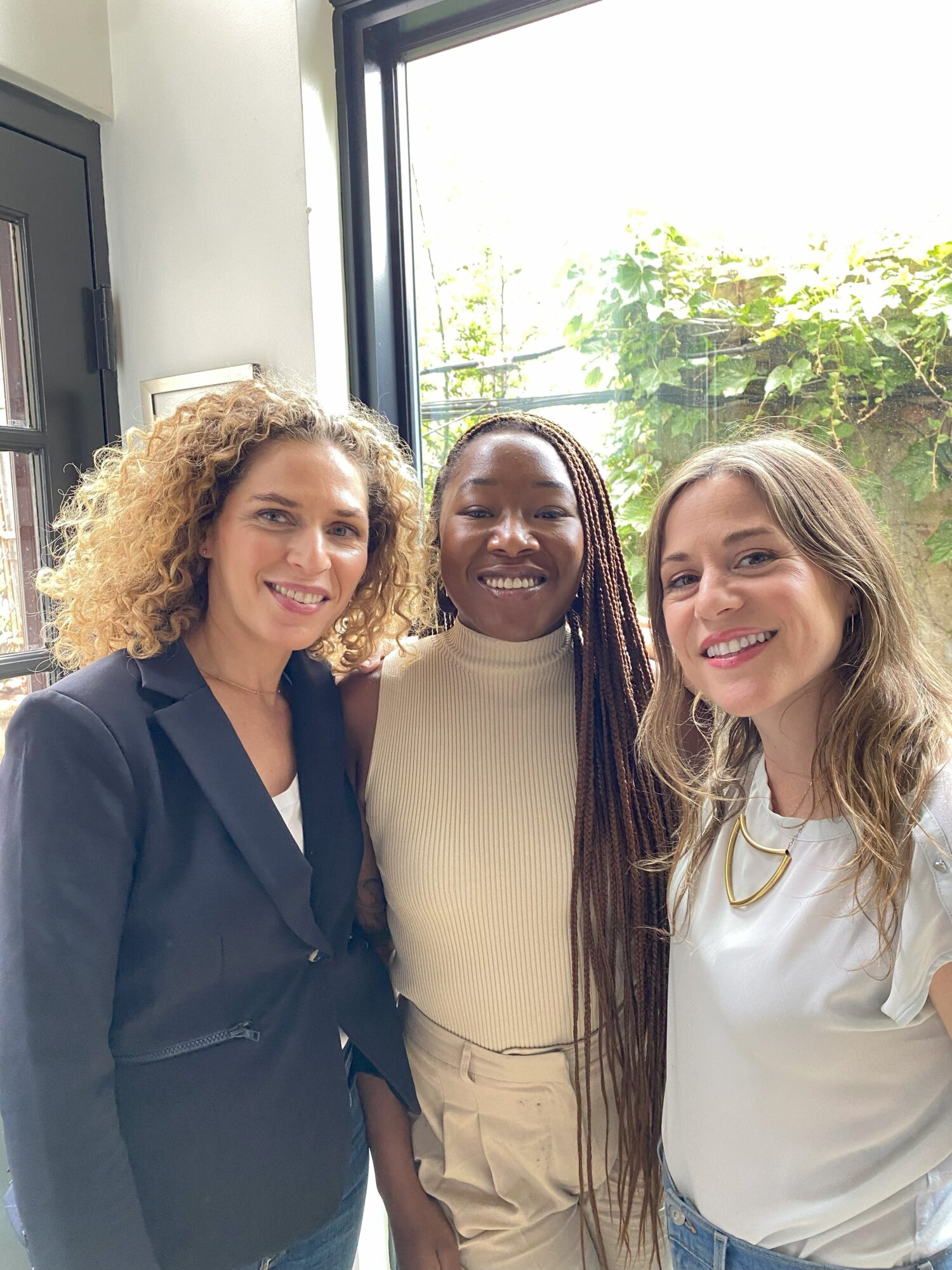 Three women smiling indoors near a window with greenery outside, posing together.