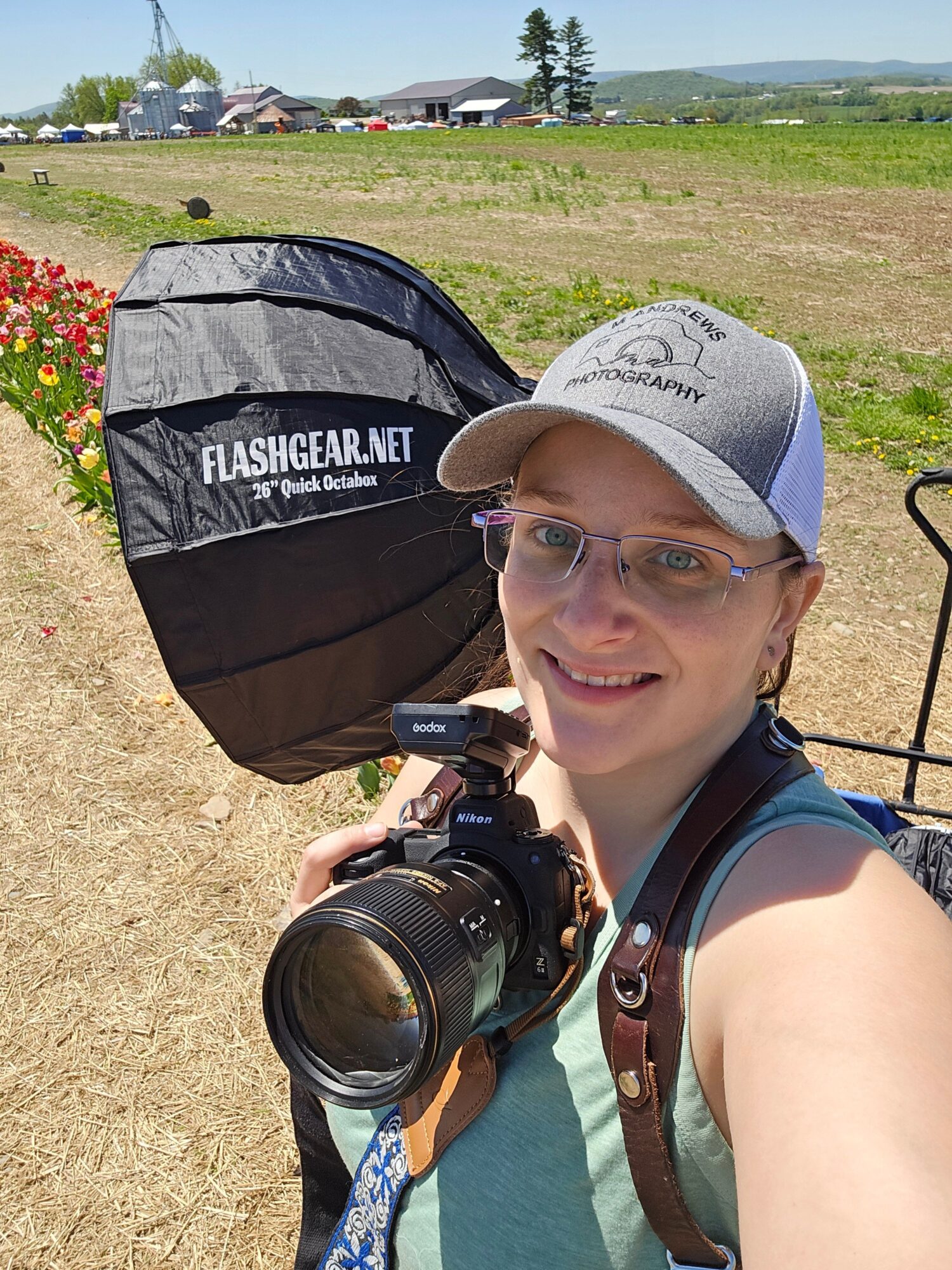 Person smiling, wearing glasses and a cap, holding a camera, outdoors with a tent and grassy area in background.