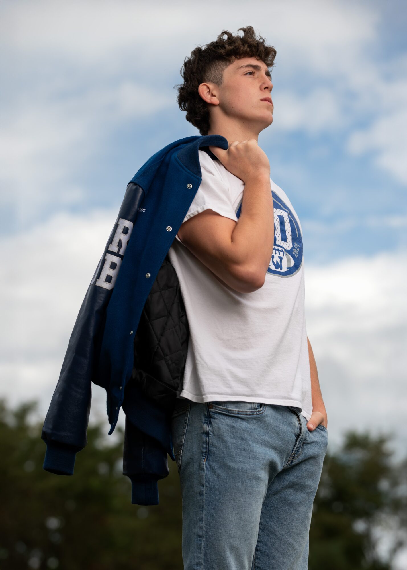 Young man standing outdoors with a jacket over his shoulder, looking up at the sky, wearing a white t-shirt and jeans.