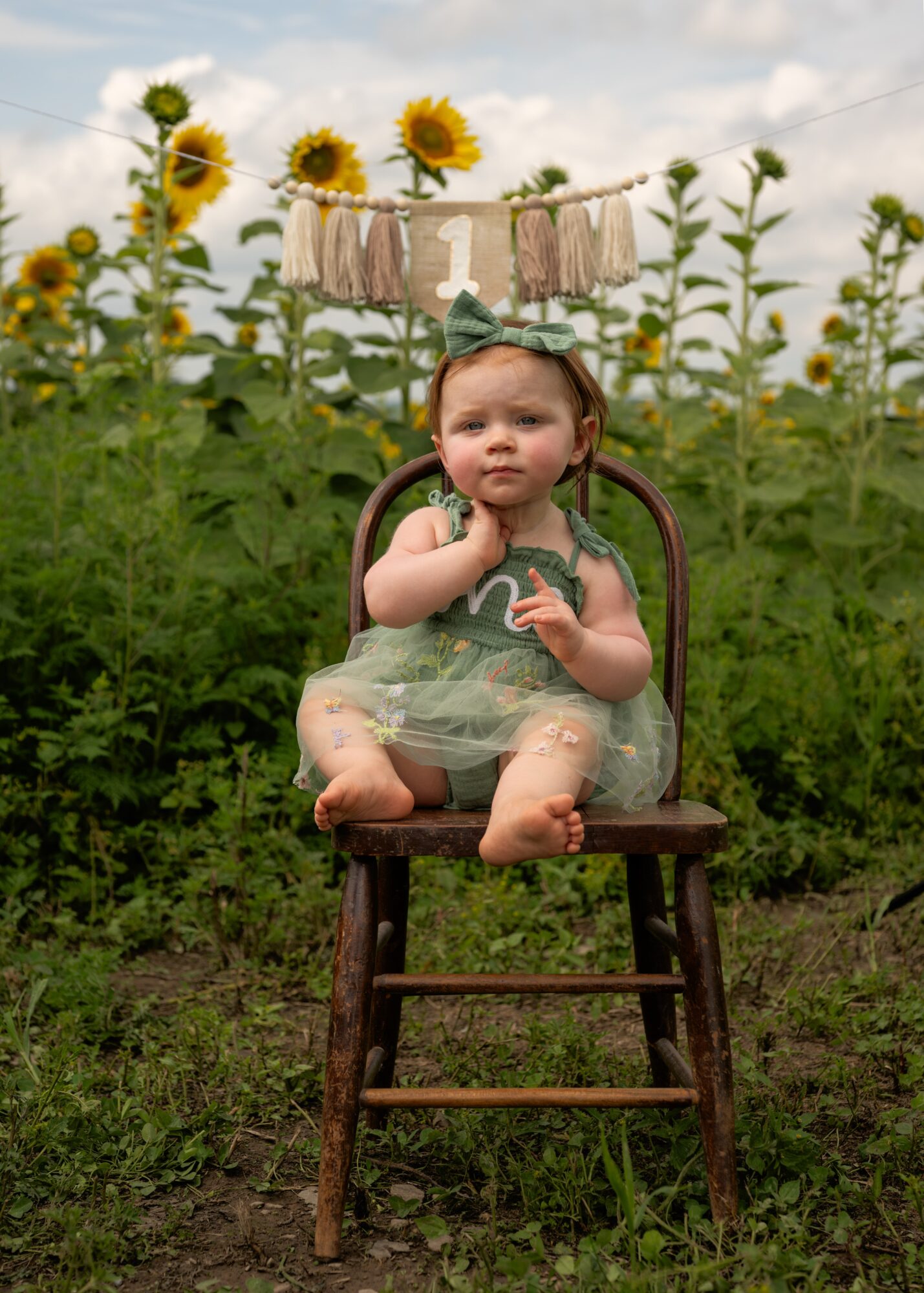 Child sitting on a wooden chair outdoors with sunflowers in the background, holding a small object, wearing a dress and headband.