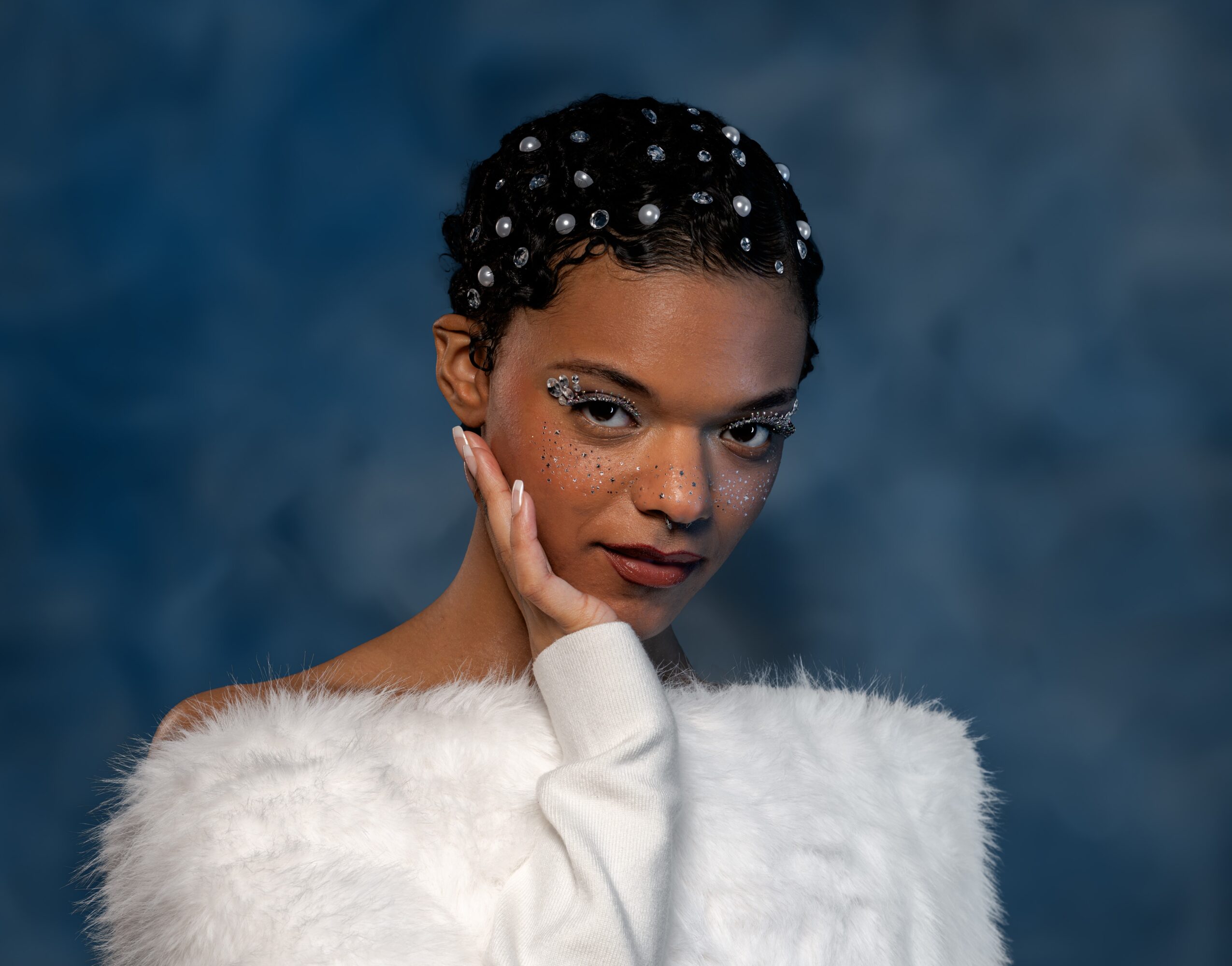 Portrait of a woman with short, curly hair decorated with pearls, wearing a white fuzzy sweater, against a dark background.