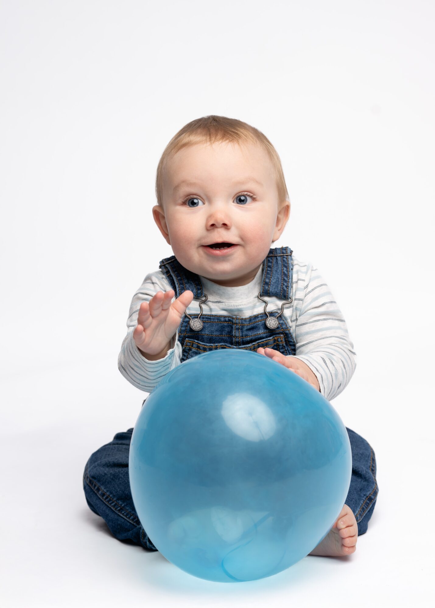 Child sitting with a large blue balloon, smiling and waving, against a white background.