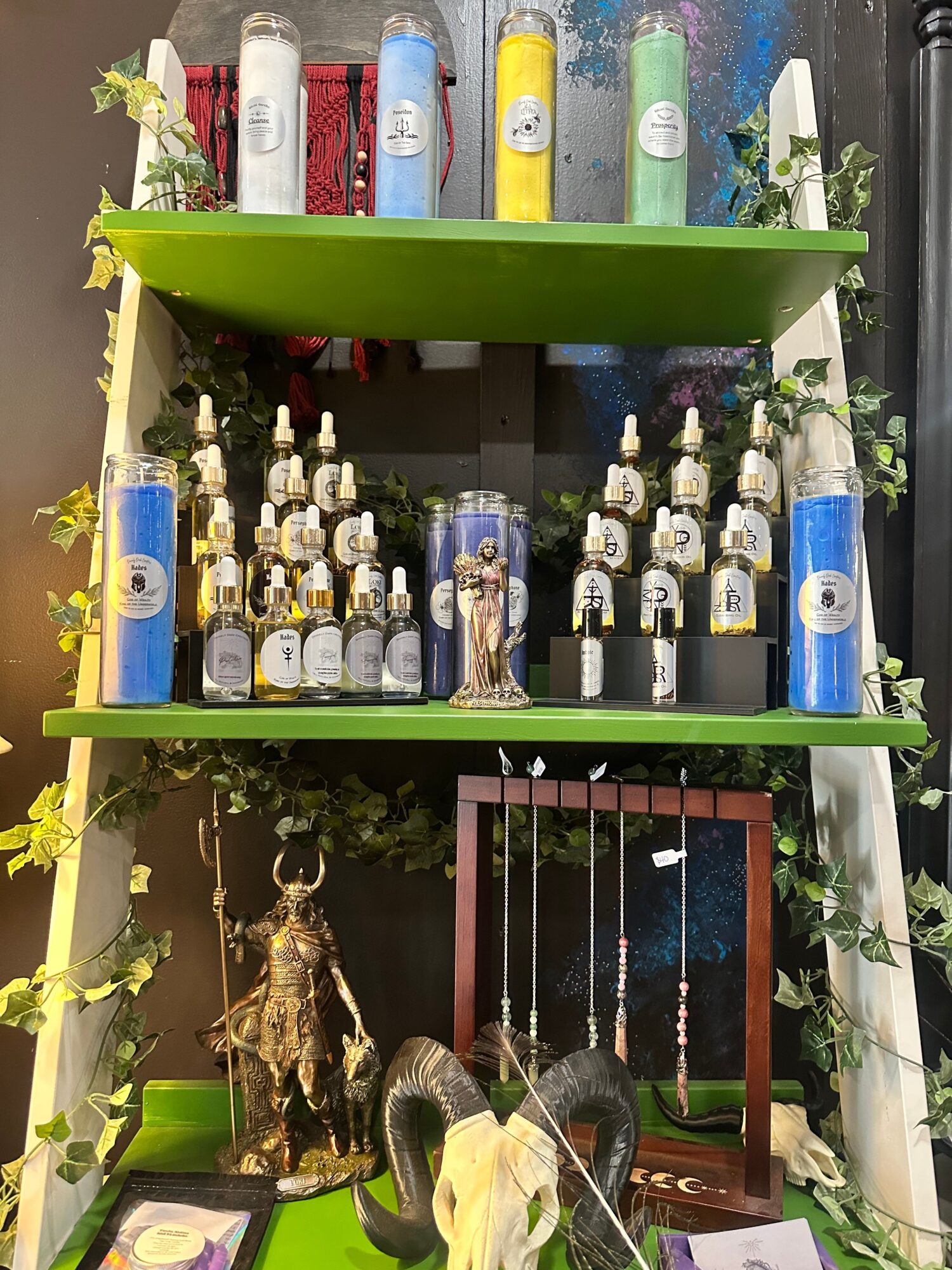 Display of various bottles and decorative items on green shelves with flowers and a framed picture, arranged in a shop or cafe.