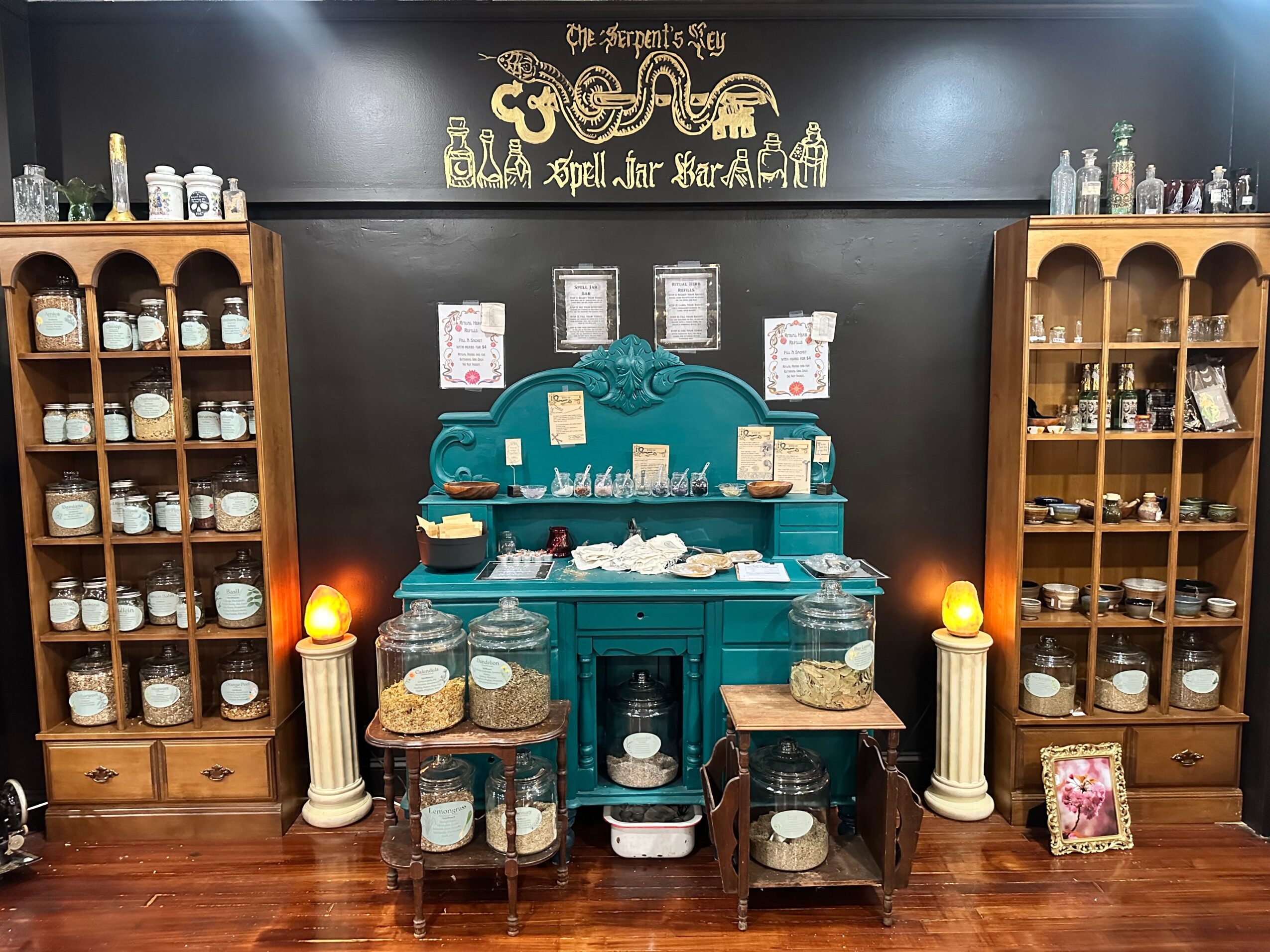 Display of jars and bottles on wooden shelves with a central teal cabinet, candles, and framed pictures on a dark wall.