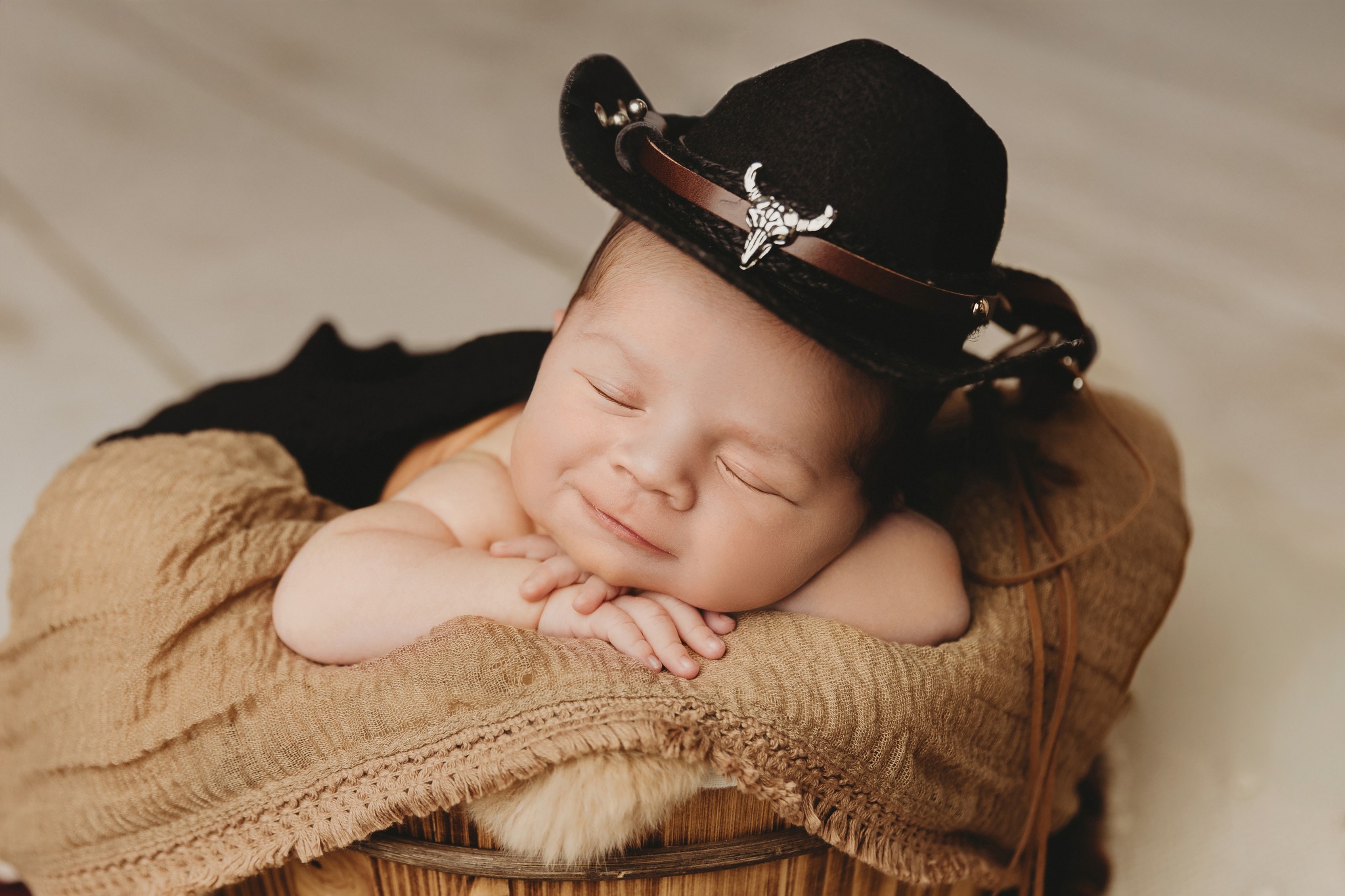 Baby with eyes closed, resting head on arms, wearing a black hat with a decorative band, lying on a blanket.