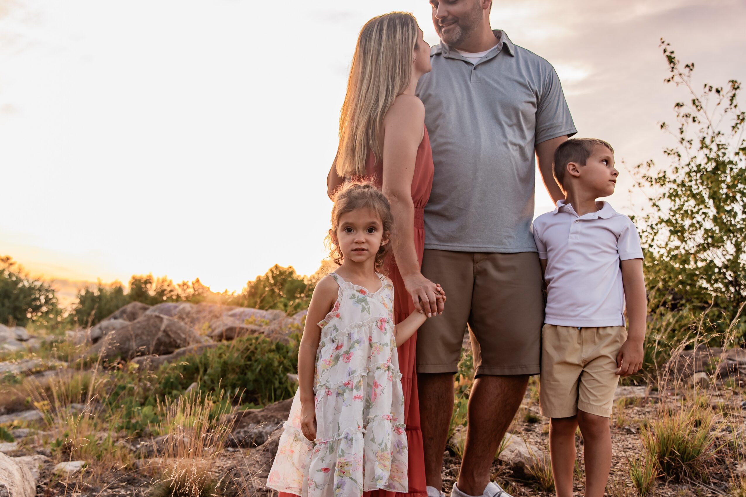 Family of four standing outdoors during sunset, holding hands, with trees and rocks in the background.