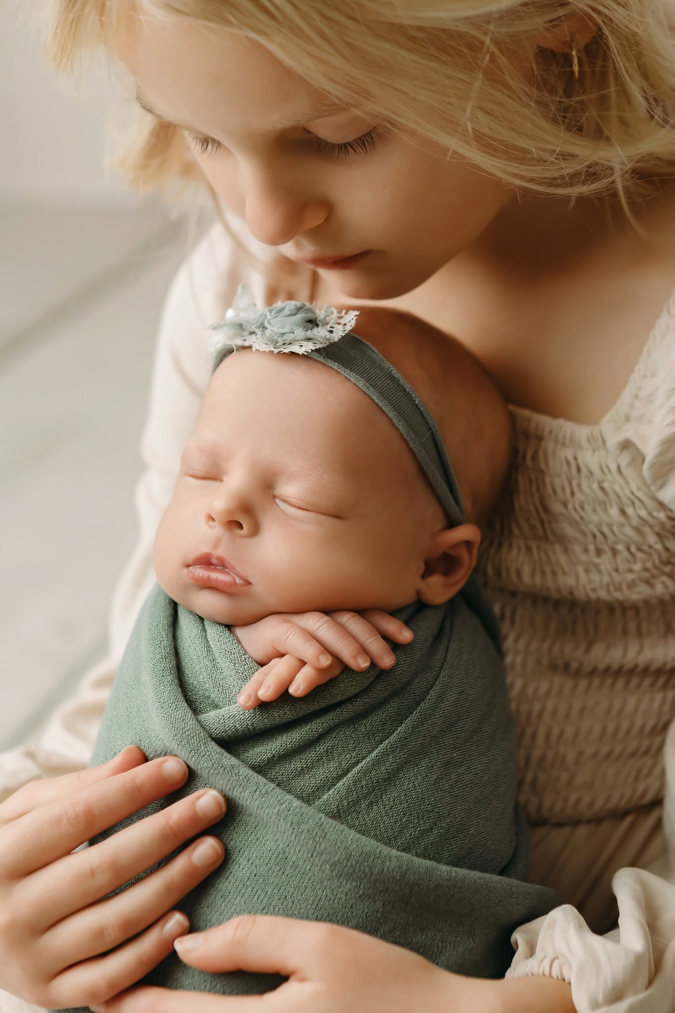Young woman gently holds a sleeping baby wrapped in a green blanket, close to her face, in soft natural light.